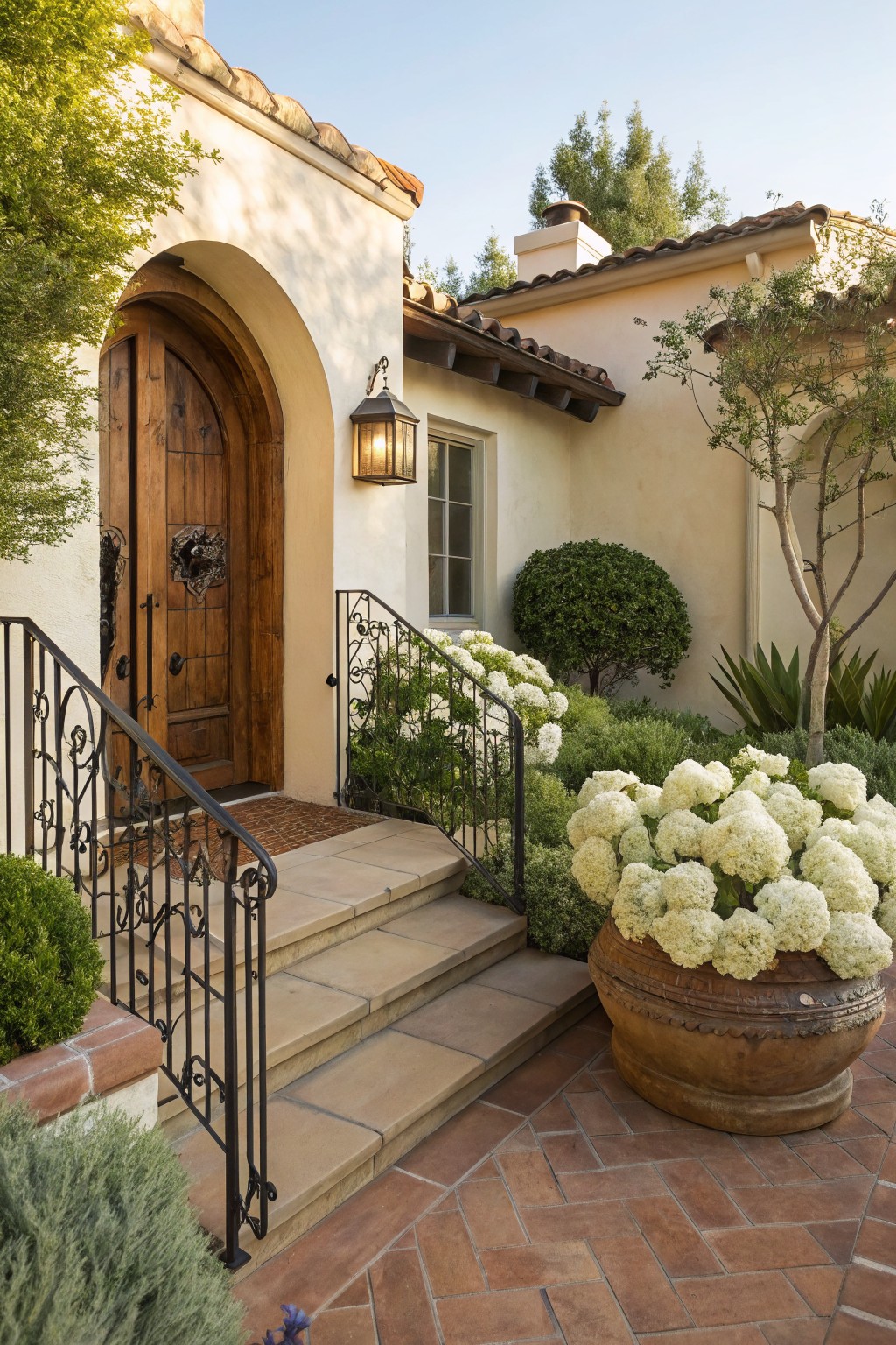 Stucco house exterior featuring an arched wooden front door, steps with black wrought iron railing, and beds of white hydrangea blooms flanked by shrubs and a large terracotta pot of hydrangeas.