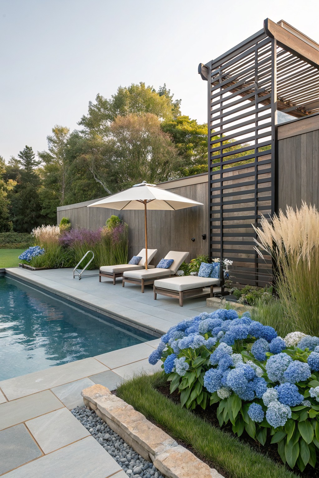 Sleek infinity-edge pool with gray stone decking and two beige chaise lounges under a white umbrella, next to a tall black slatted fence screen on a wood-clad wall, bordered by dense blue hydrangea bushes, pinkish ornamental grasses, and low groundcover.