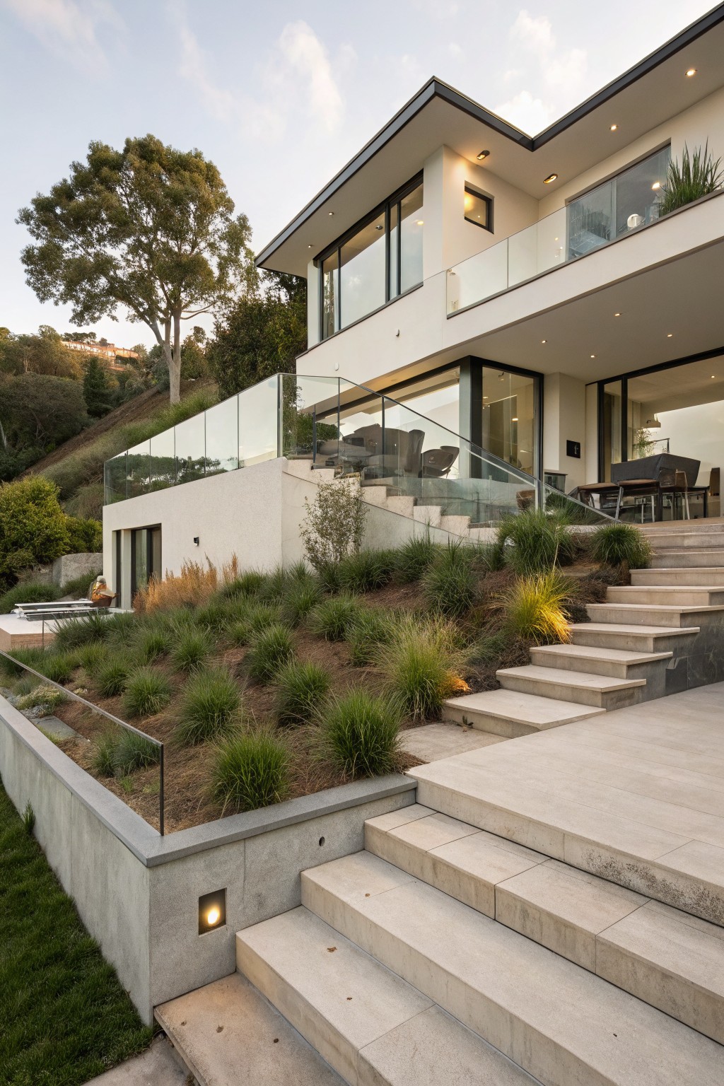 White modern house on a hillside with wide concrete terraced steps, glass railings, balcony overlooking landscaped grasses and a lower patio area.