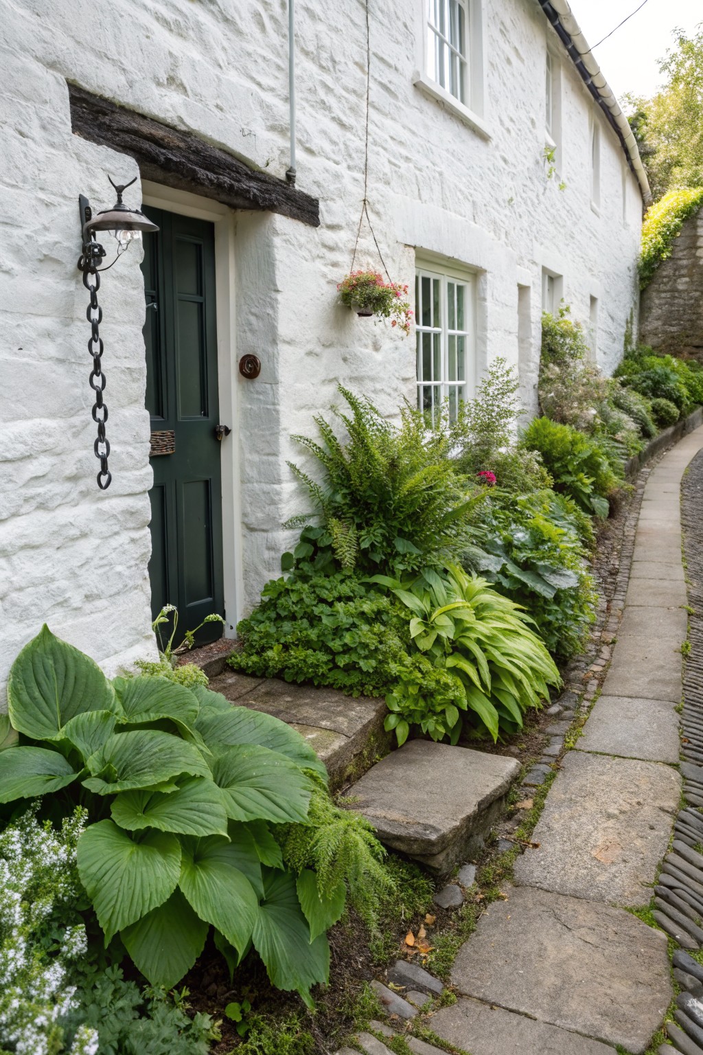 White stone cottage with dark green front door and lantern, bordered by dense green plantings of hostas, ferns, and flowering shrubs along a curving stone path.