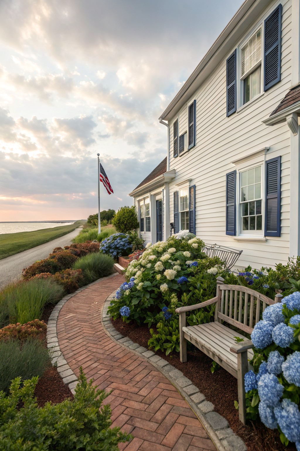 White clapboard house with blue shutters and an American flag pole, viewed from a curved red brick pathway edged in blue and white hydrangea bushes with a wooden bench beside a body of water.