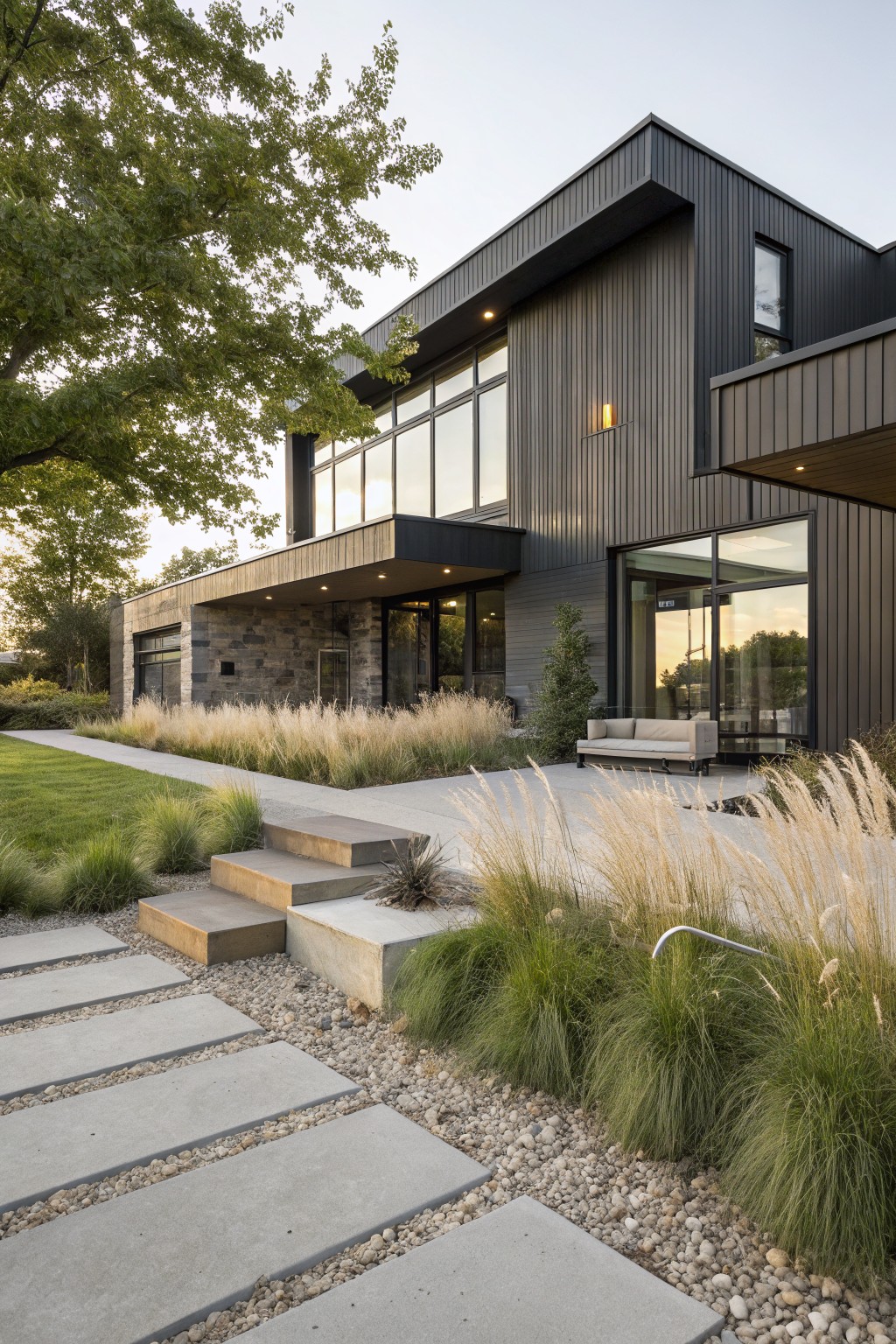 Modern black-clad house exterior with large glass windows and stone garage, concrete paver pathway edged by tall ornamental grasses with white plumes leading to entry steps, lawn and trees in background at dusk.