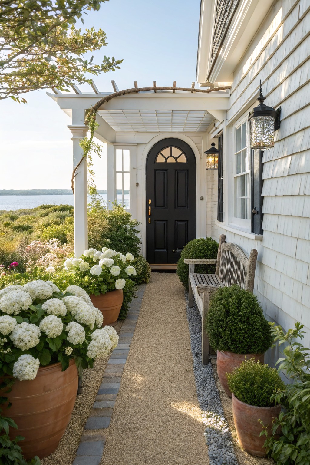 Shingled white house exterior with arched black front door under a pergola, gravel path lined with terracotta pots of white hydrangeas and boxwoods, wooden bench, and view of bay in background.