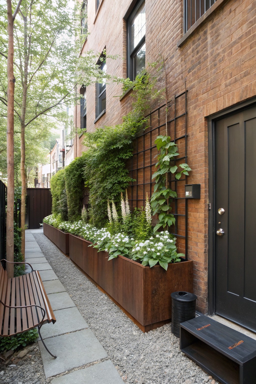 Narrow brick pathway lined with long rectangular corten steel planters filled with white flowers and green plants, wooden bench nearby, black entry door, and climbing vines on metal trellis against brick wall.