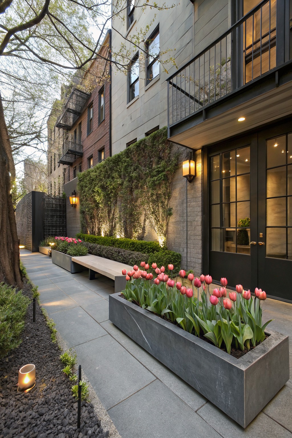 Stone pathway bordered by large rectangular concrete planters filled with red tulips, a wooden bench, green hedges on walls, lanterns, and black-framed glass entry doors on brick and stucco buildings.
