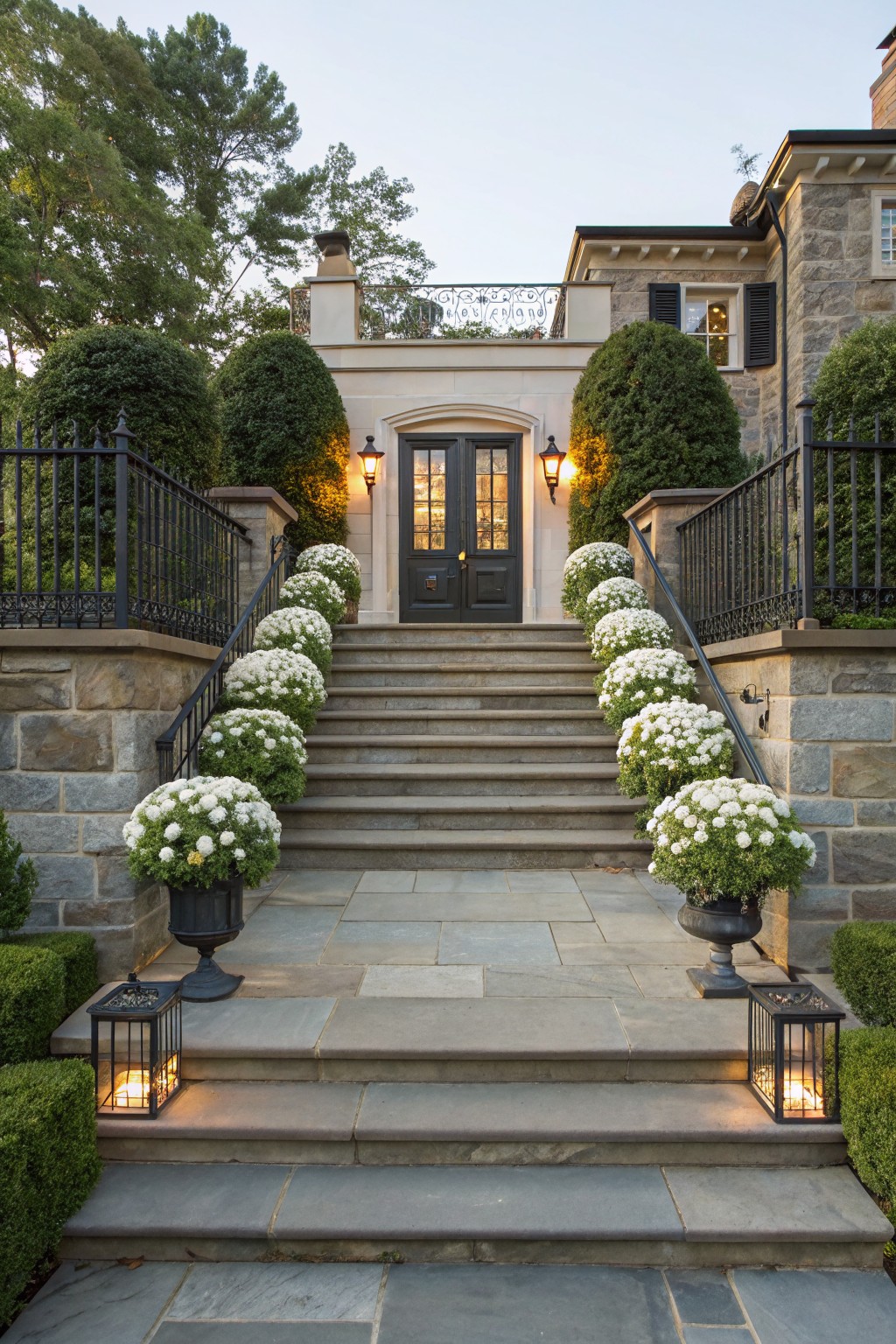 Stone steps leading to a dark wood double door entrance, lined with pots of white spherical flowers along iron railings on both sides, with black lanterns at the base and topiary shrubs nearby.