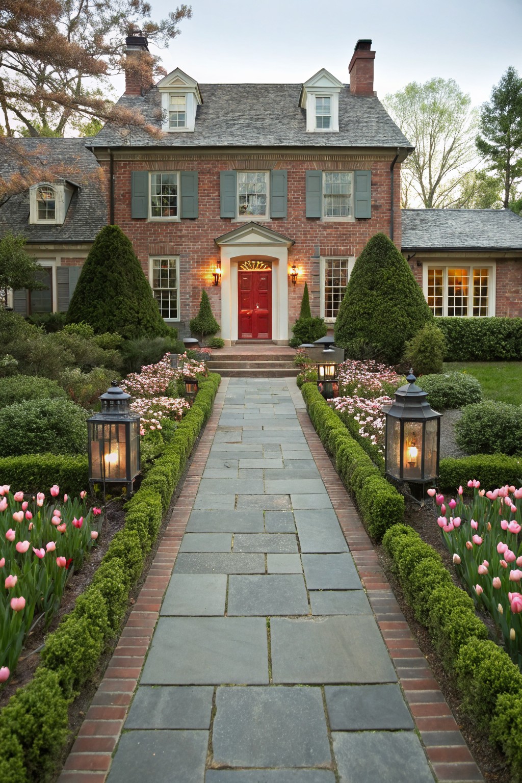 Red brick house with slate stone pathway lined by boxwood hedges and pink tulip flower beds on both sides, lanterns along the path, leading to a red front door flanked by evergreens.