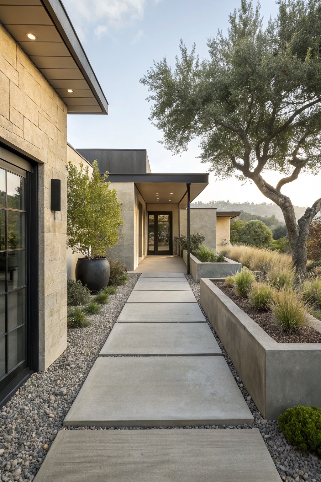 Contemporary house exterior featuring a pathway of large rectangular gray concrete slabs in gravel leading to a glass-fronted entry, bordered by a raised concrete planter with ornamental grasses, stone walls, potted trees, and oak trees with hills in the background.