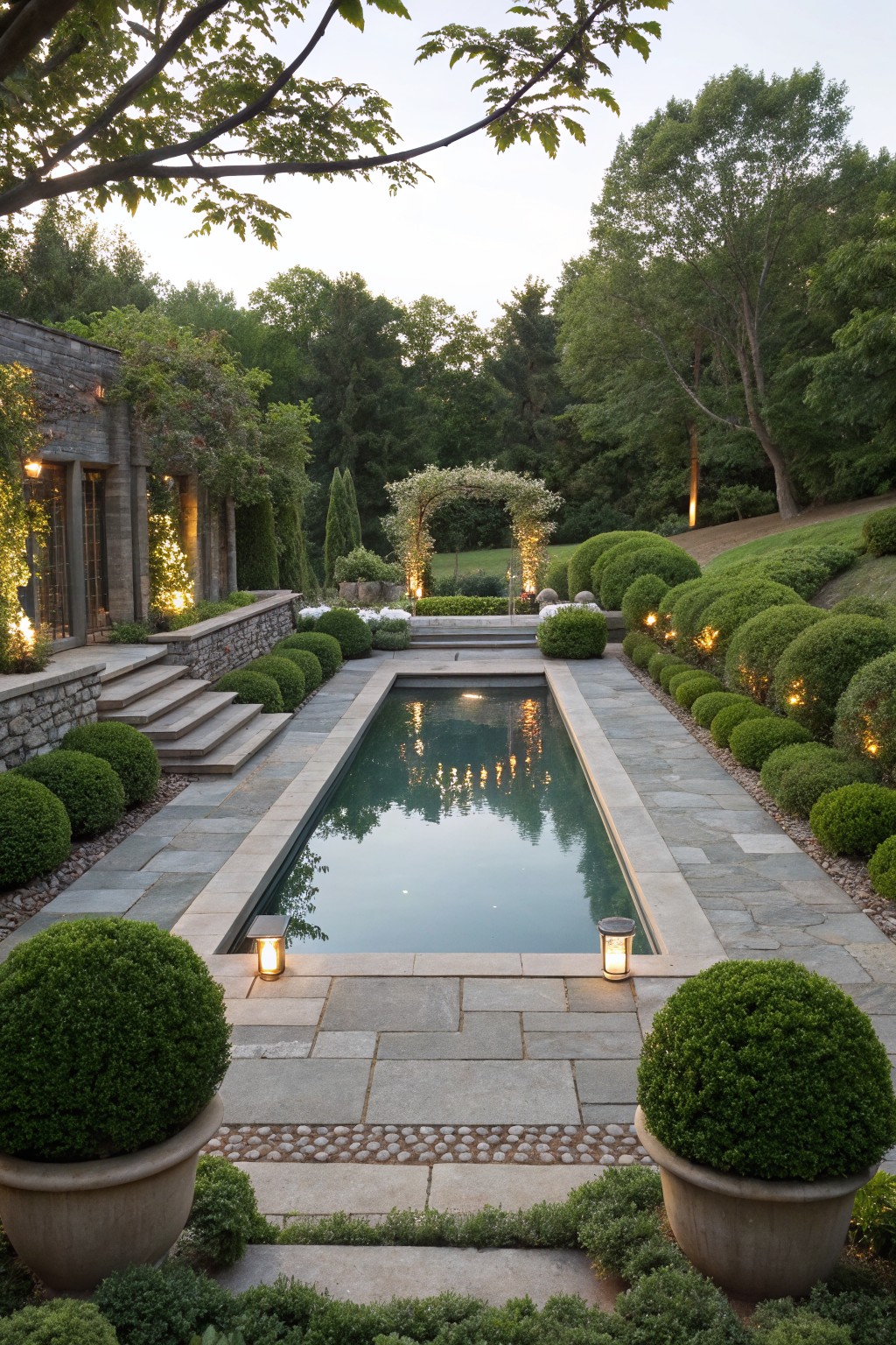 Rectangular pool with gray flagstone surround and pebble borders, flanked by round boxwood shrubs, stone retaining walls with steps, and landscape lanterns in a wooded backyard.