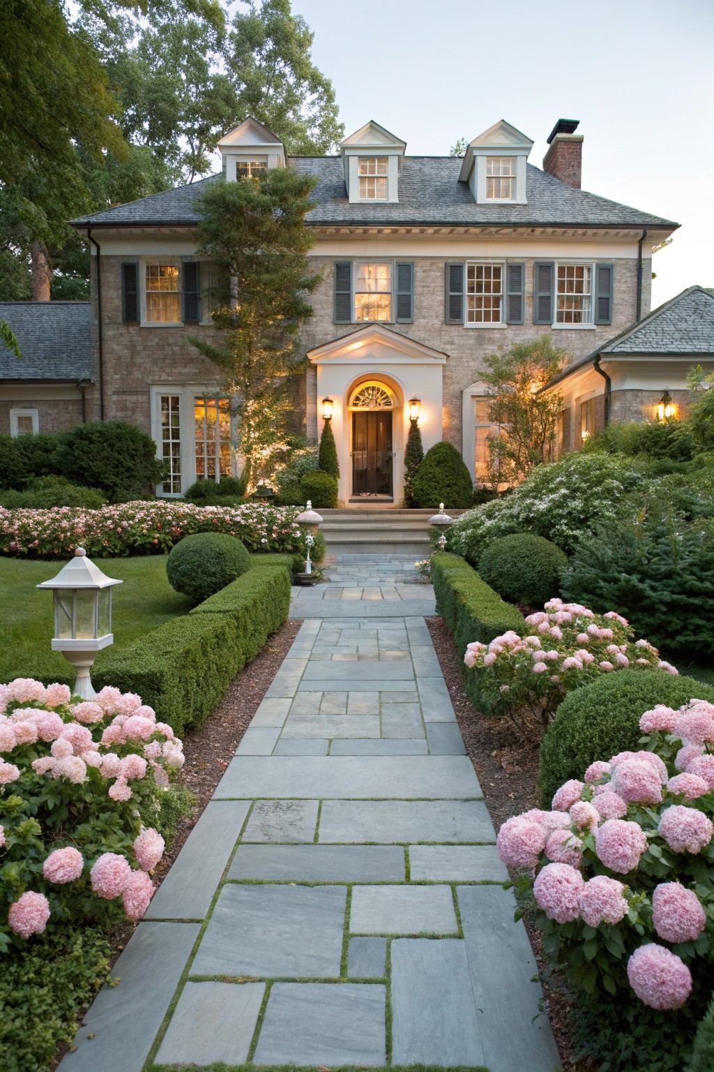 Stone slab pathway bordered by large pink hydrangea bushes and boxwood hedges leading to the columned entrance of a brick house with shuttered windows and landscape lighting.