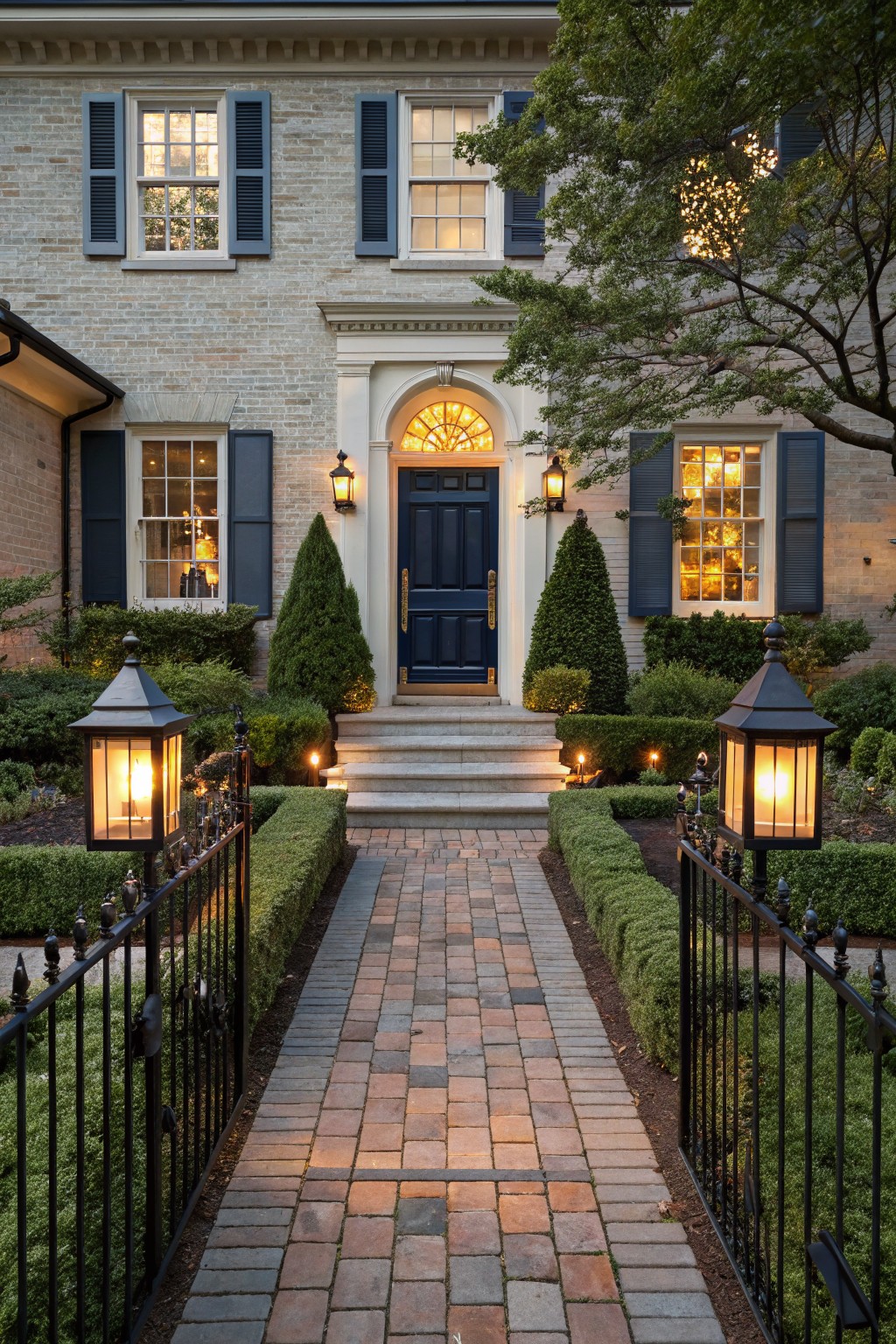 Evening photo of a beige brick house with navy front door, brick pathway lined by black iron lanterns and boxwood hedges through a gated entry.