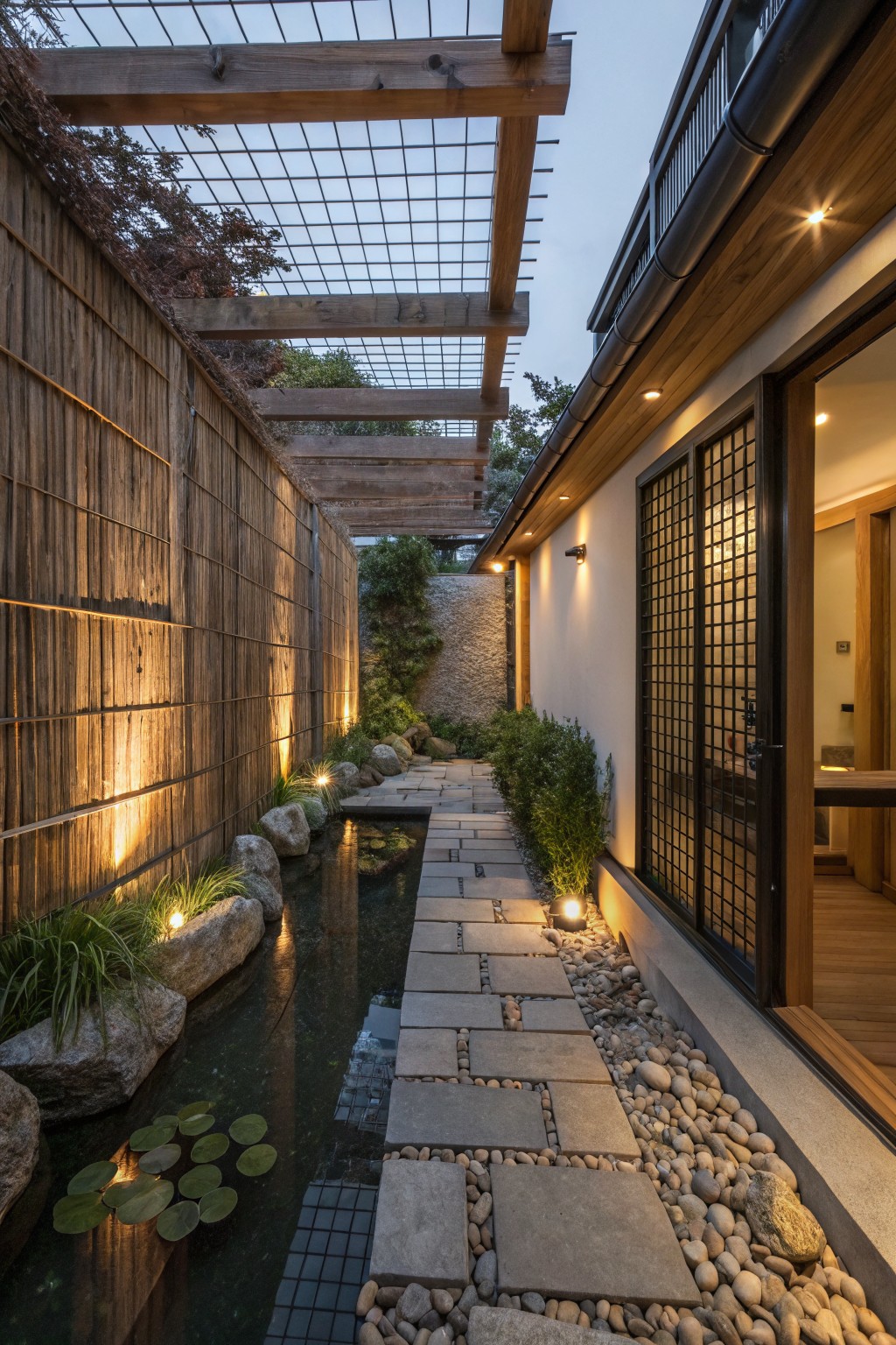 Narrow garden pathway lined with tall bamboo fences on one side and house exterior on the other, featuring stone pavers, small pond with lily pads and rocks, wooden pergola overhead, and warm exterior lighting.