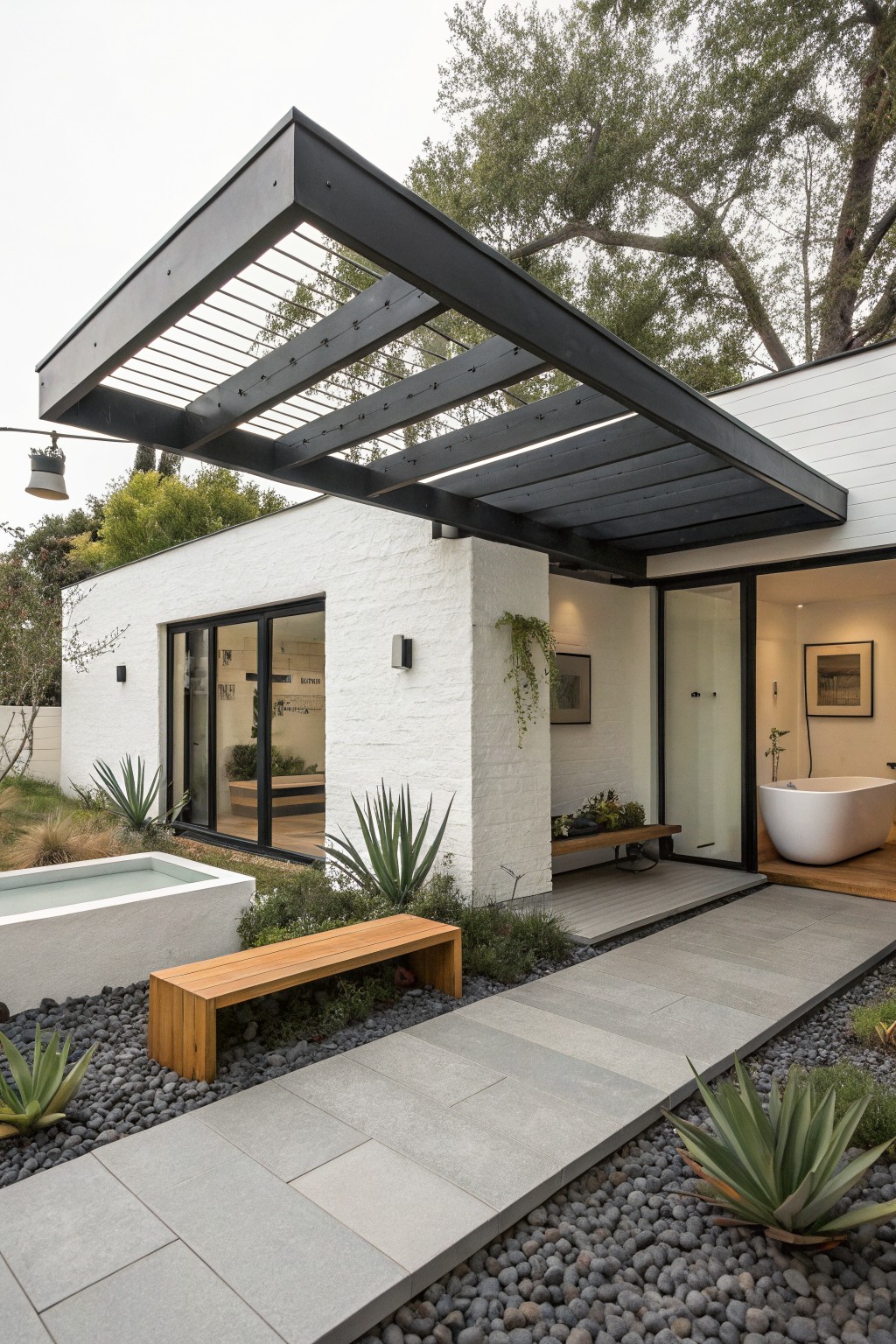 White stucco house exterior with black metal grate pergola canopy over gray slate entry path lined by agave plants and black gravel, wooden bench alongside, small rectangular pool nearby.
