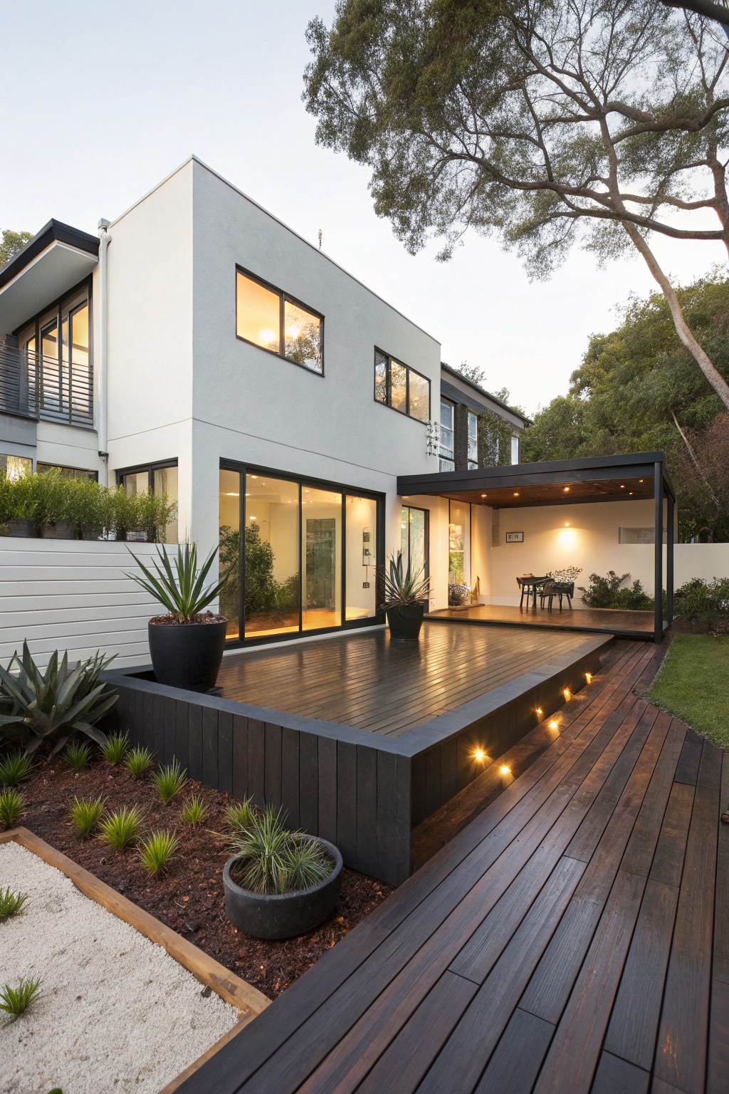 Modern white two-story house with large glass doors and windows opening to a raised dark timber deck platform, edged with gravel beds, agave plants, and potted greenery, featuring low-level lighting along the deck at dusk.