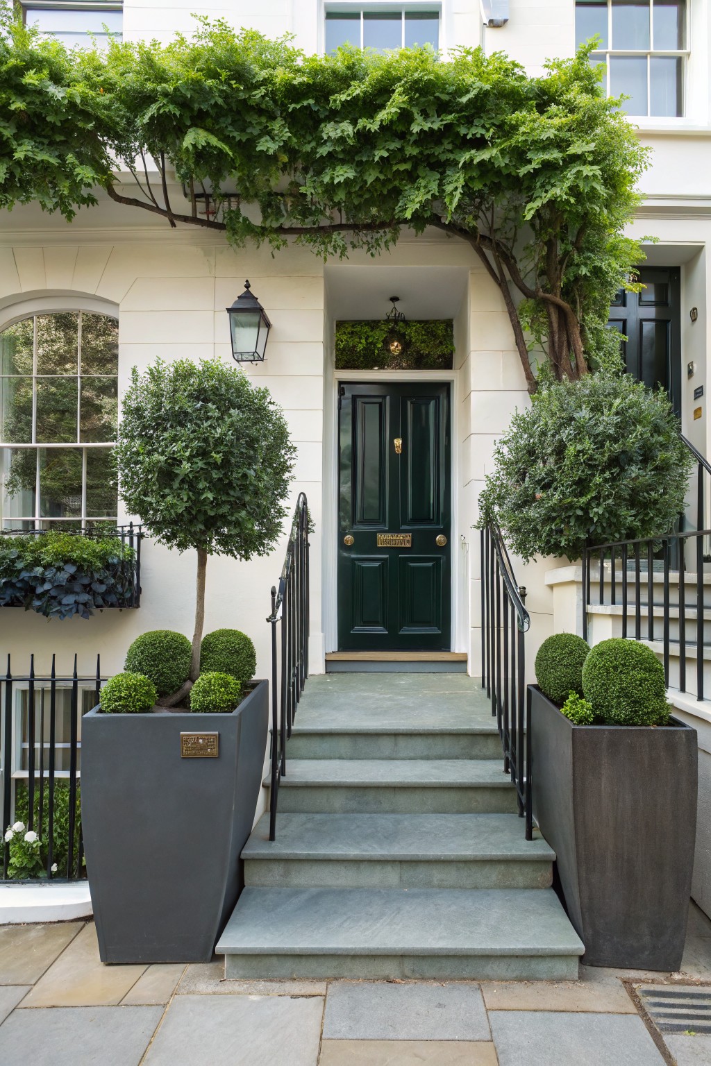 White stucco house facade with dark green front door, flanked by stone steps with black iron railings and large gray cylindrical planters holding spherical topiary trees and shrubs, ivy vine climbing above the entry.