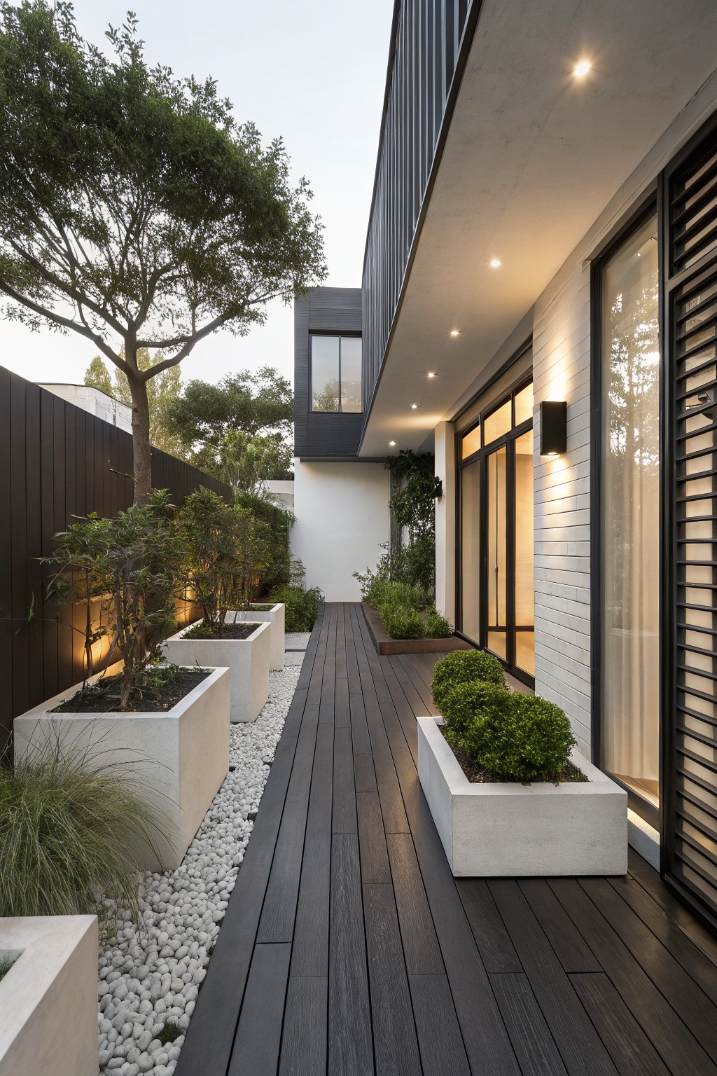 Side view of a modern house with a dark wood deck pathway bordered by white concrete planters holding greenery and shrubs, a pebble strip, grasses, and trees against white and black architecture in evening light.