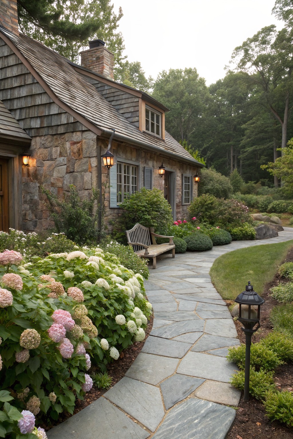 Rustic shingled stone cottage with front door, flanked by windows and lanterns, leading to a curved flagstone path bordered by blooming pink and white hydrangeas, a wooden bench, and surrounding shrubs and trees.