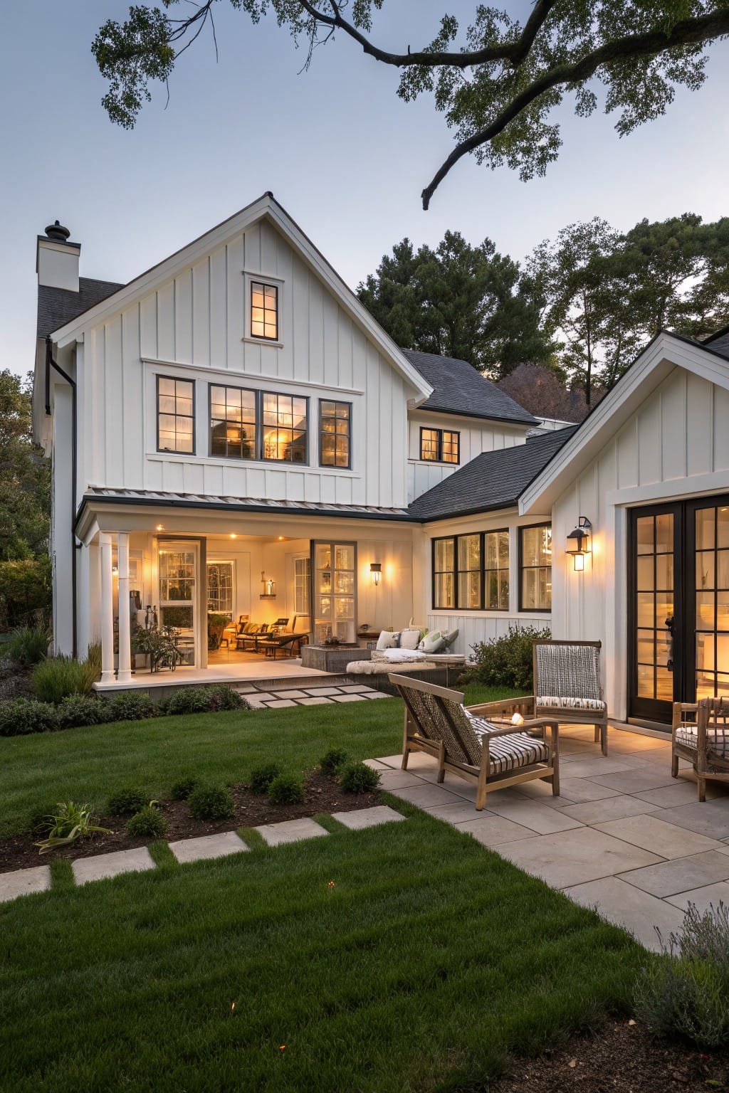 White board-and-batten farmhouse exterior at dusk with covered patio, folding glass doors open to lawn, paver path, Adirondack chairs, and surrounding trees.