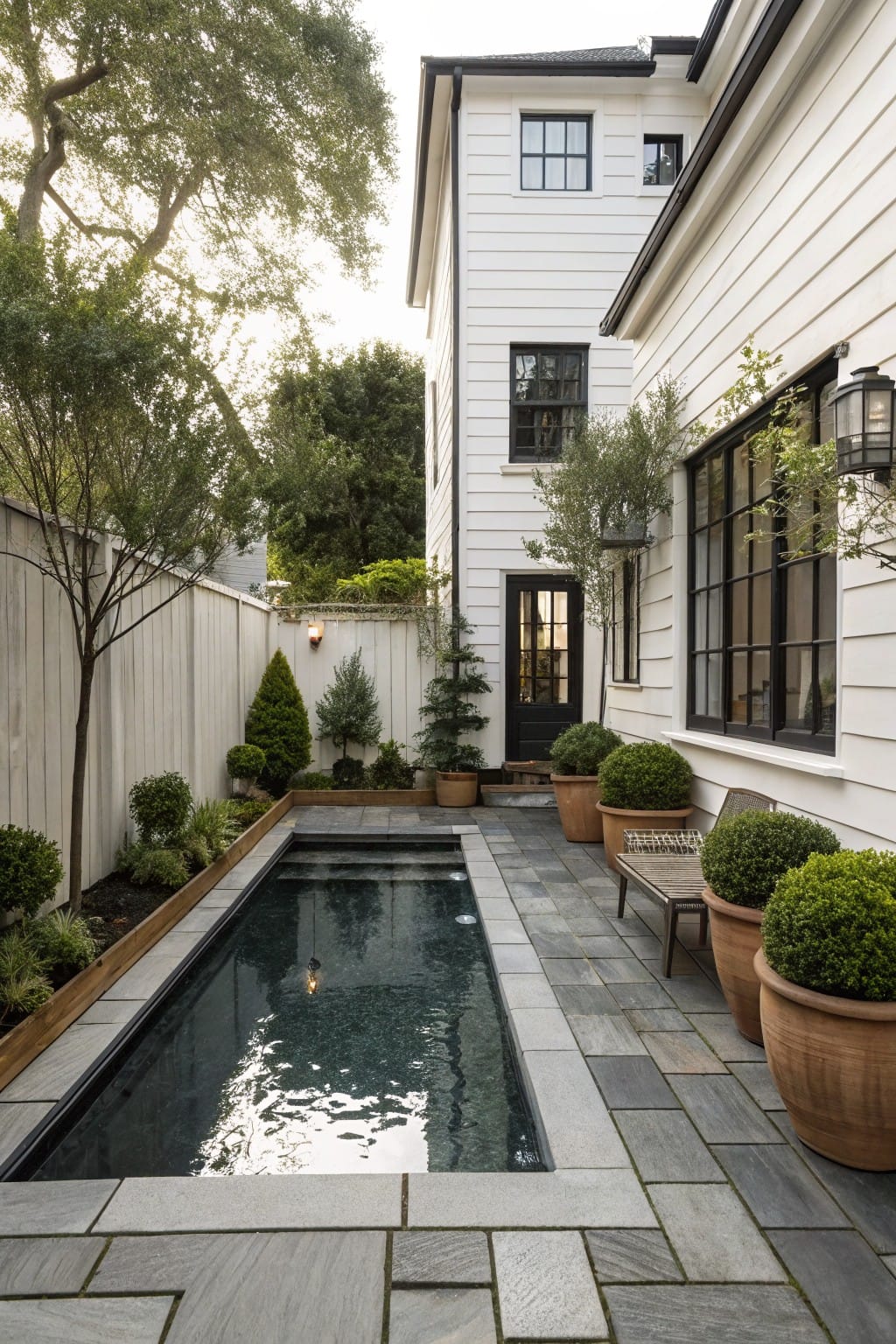 White clapboard house with black-framed windows next to a narrow rectangular in-ground pool paved in gray stone slabs, with large terracotta pots of greenery and a wooden fence along the side yard.