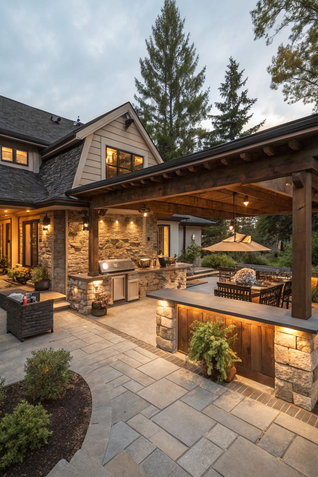 Backyard patio with covered stone outdoor kitchen under wood beams, built-in grill and counters, paver flooring, seating, plants, and house in background.