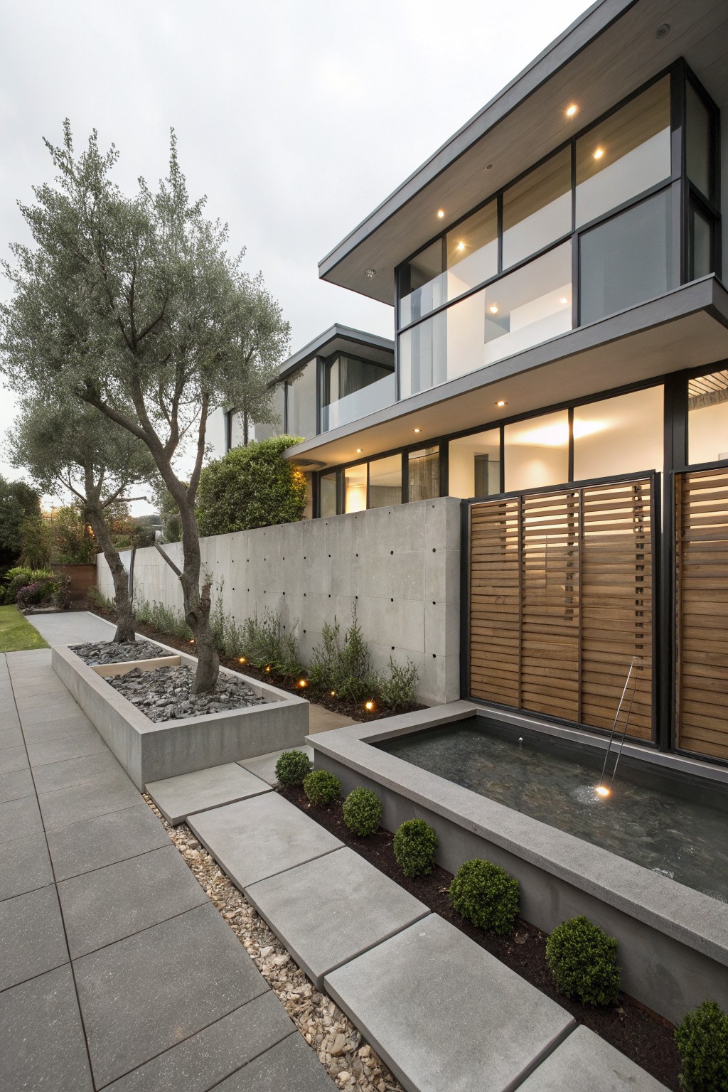 Modern house exterior with tall slatted wooden fence on concrete wall, olive trees in rectangular planters, boxwood shrubs, small reflecting pool with fountain, and gray paver pathway.