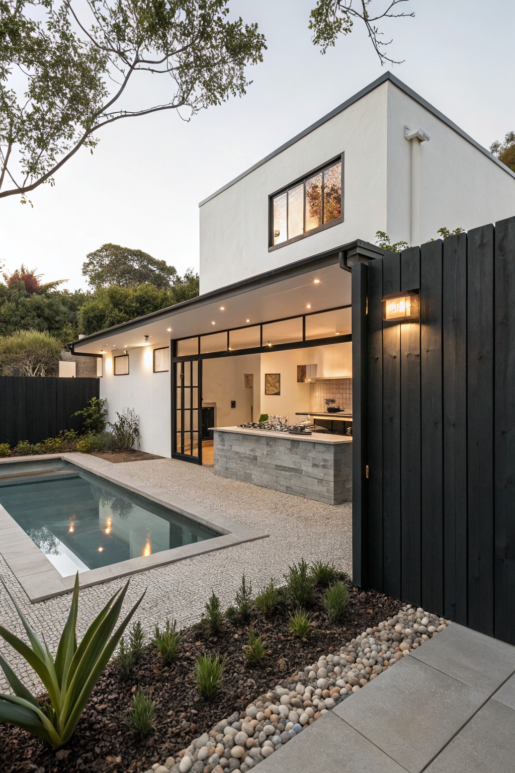 White modern house exterior with black vertical slat timber fence enclosing an outdoor kitchen, stone countertop bar, infinity pool, agave plants, and pebble ground cover.