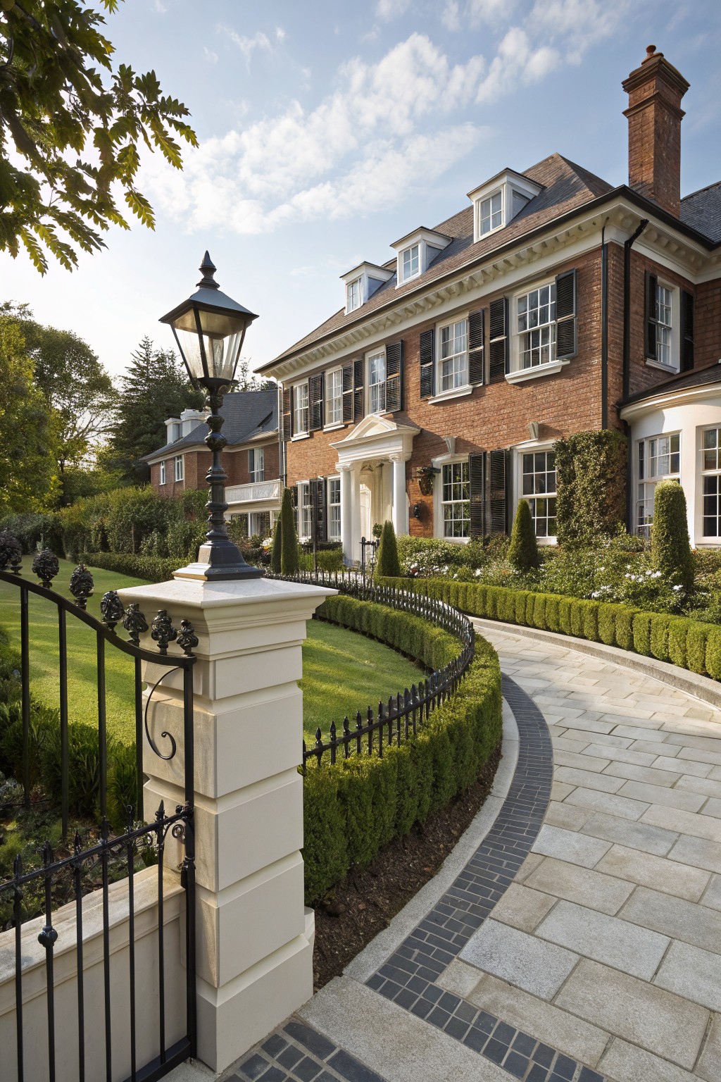 Red brick house with white shutters and dormer windows behind a curved black wrought iron fence on stone pillars, edged by low boxwood hedges along a paved pathway, with a lantern light post at the entrance.