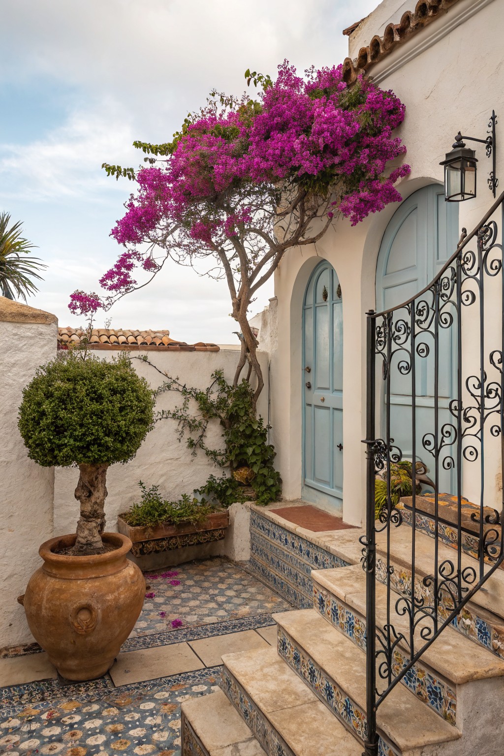 White stucco courtyard entrance with a black wrought iron gate on tiled stone steps leading to two arched blue doors, purple bougainvillea tree against the wall, terracotta pot with tree, boxwood shrub, and colorful tile flooring.
