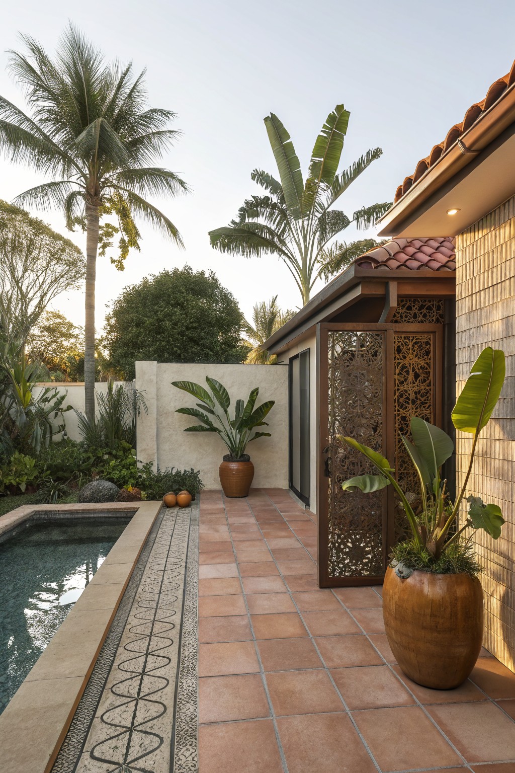 Outdoor pool with stone coping and mosaic tile border adjacent to terracotta-tiled patio, large potted plants, intricate metal lattice screen on a door and wall next to a brick house, surrounded by palm trees and banana plants.