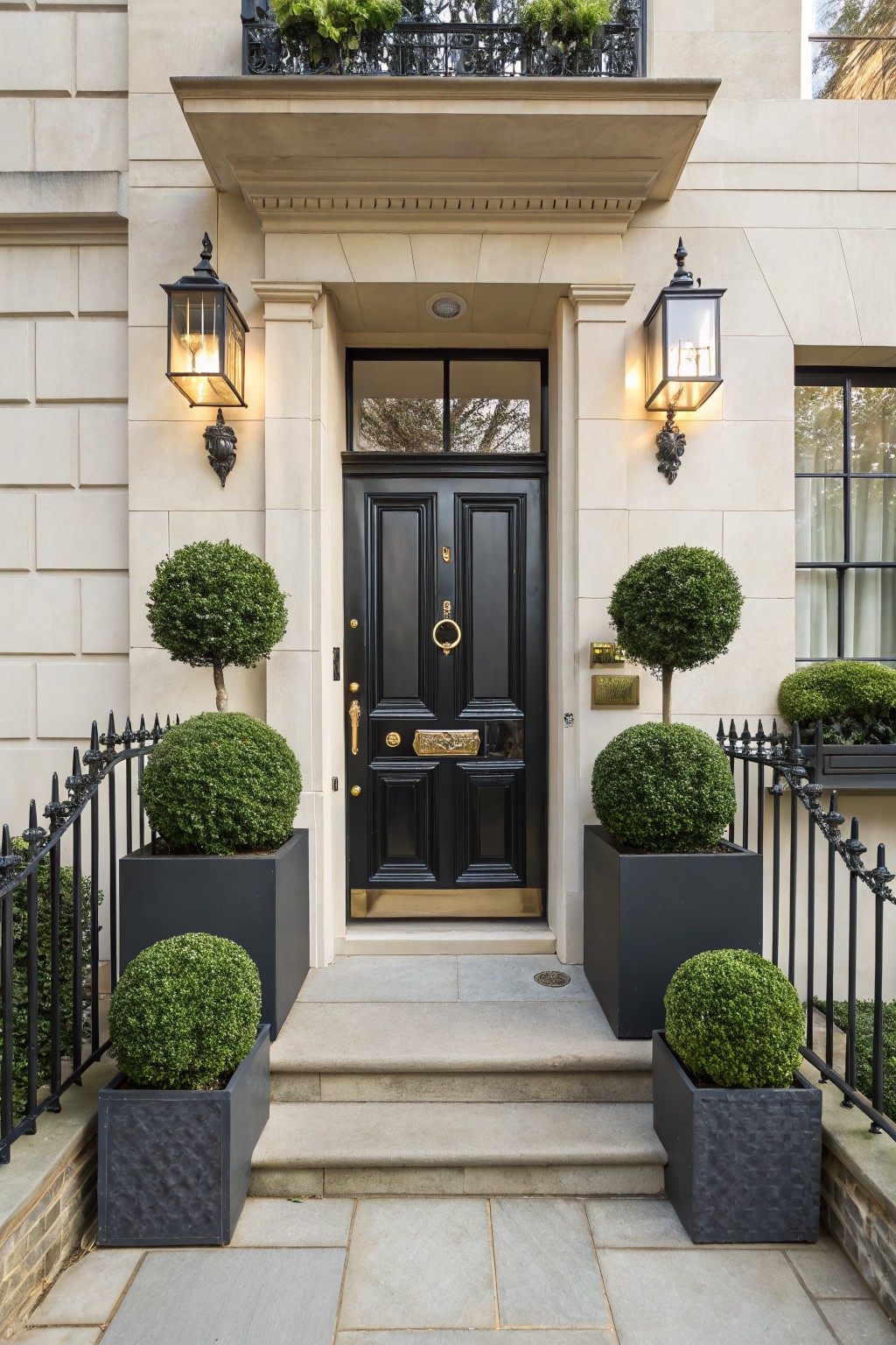 Beige stone townhouse entrance with black double front door, gold knocker and letterbox, flanked by topiary boxwood plants in black square pots on stone steps, wrought iron railing, and wall lanterns.
