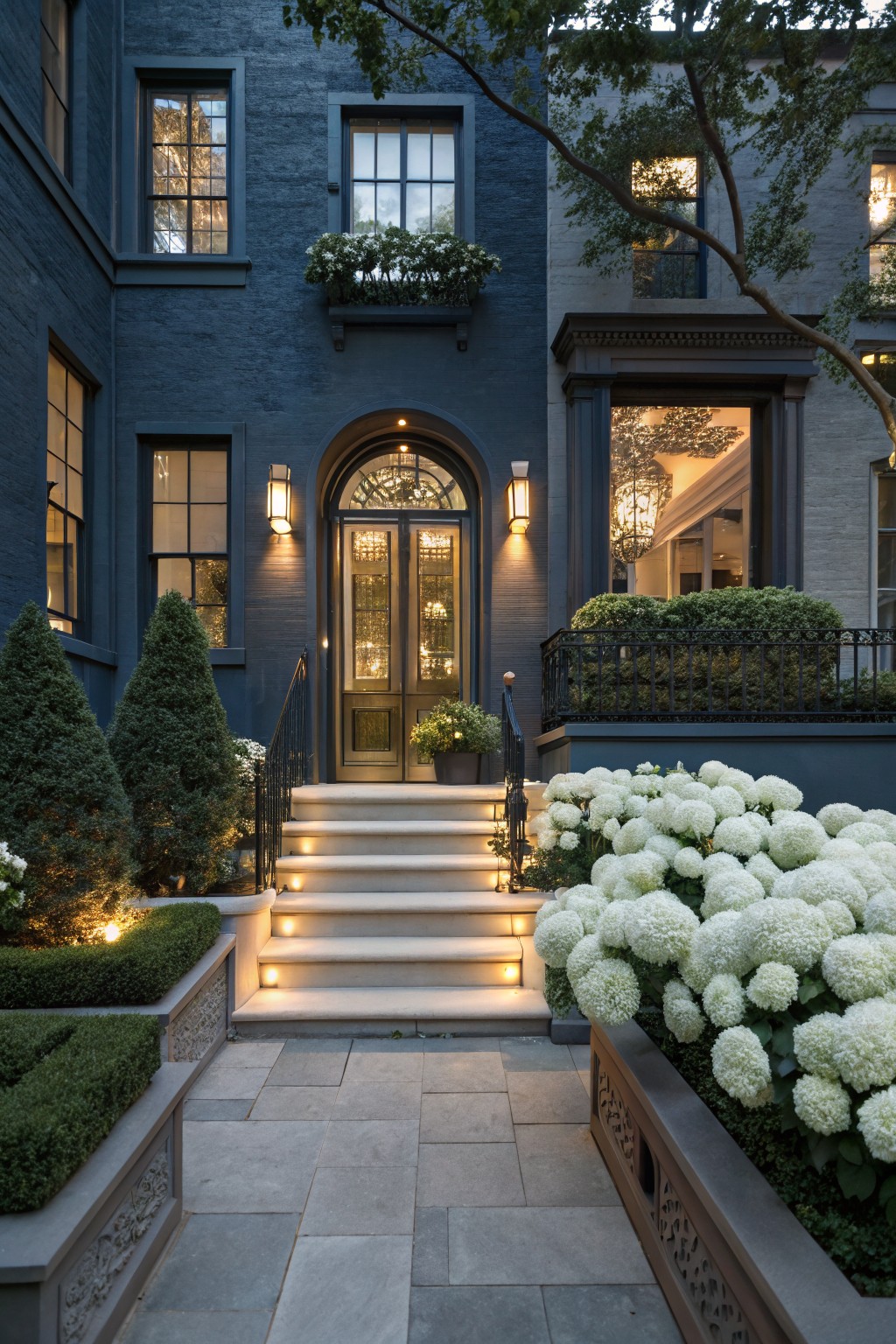 Dark navy brick townhouse facade with arched glass entry doors, lit stone steps flanked by low boxwood hedges and large white hydrangea flower beds in stone planters, leading to a paved walkway at dusk.