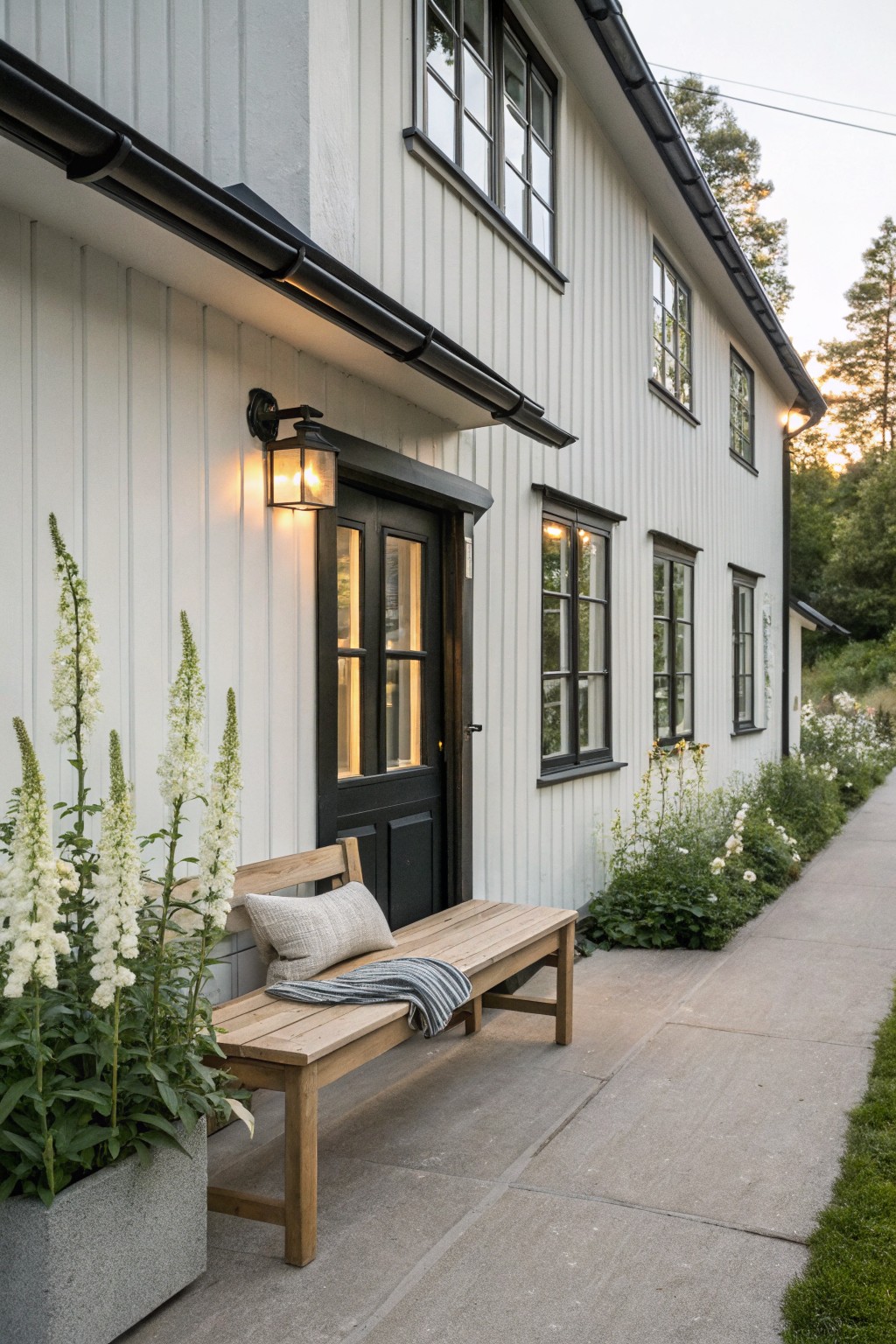 White board-and-batten house exterior with black double door, lit lantern, wooden bench with pillows and throw, tall white foxglove plants in gray pot and garden beds along concrete path flanked by greenery.
