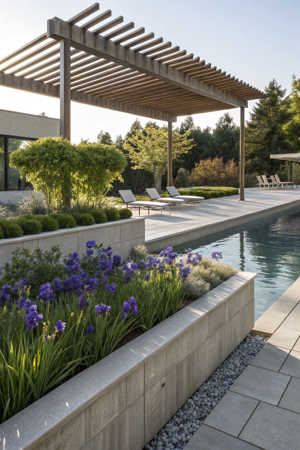 Modern infinity-edge pool with purple irises and grasses in raised concrete planters along the edge, wooden pergola with lounge chairs nearby, stone decking, and trees in the background.
