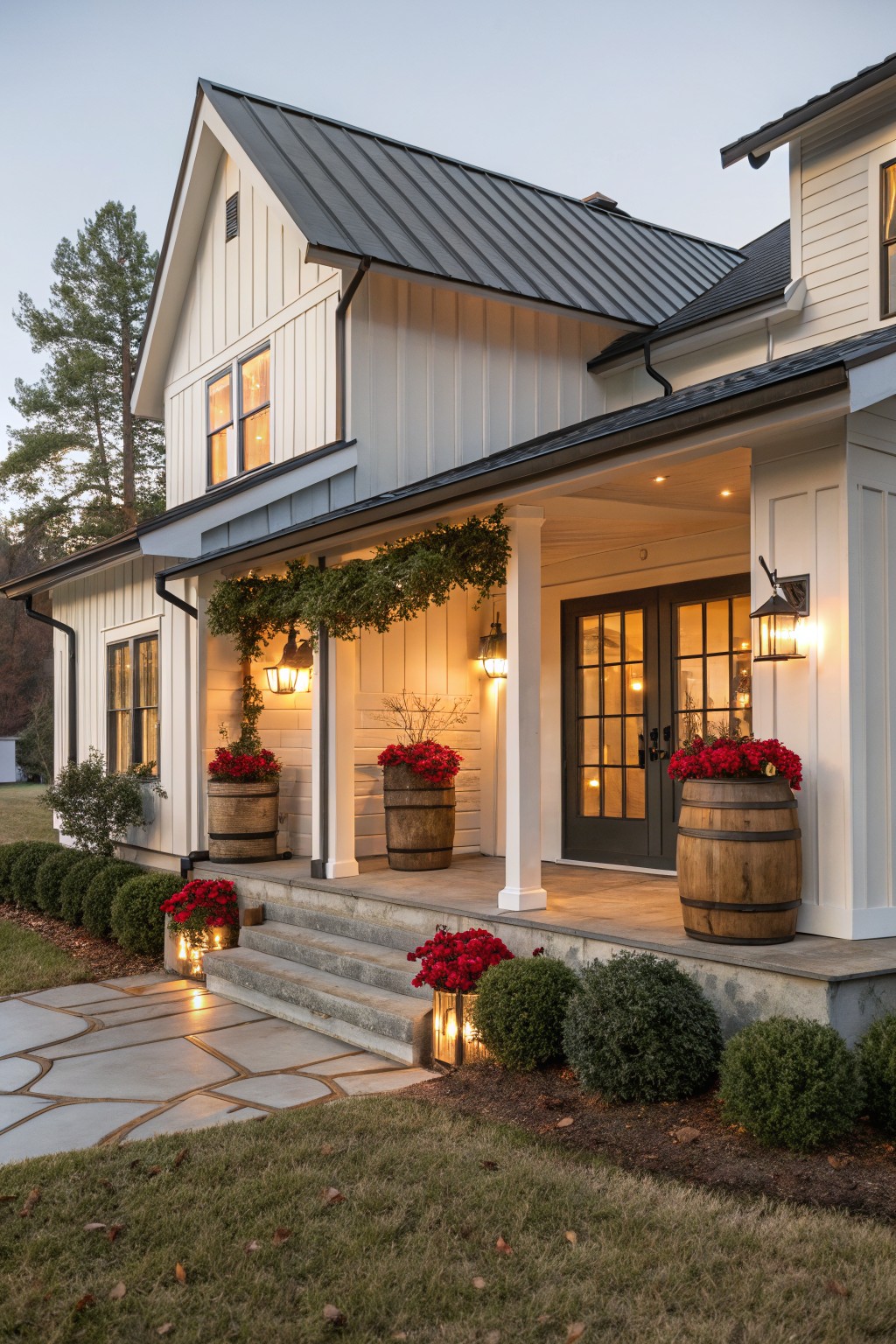 White board-and-batten house with dark metal roof and covered porch, featuring whiskey barrel planters filled with red poinsettias flanking black double doors, garland on porch columns, lanterns, and lit stone pathway at evening.