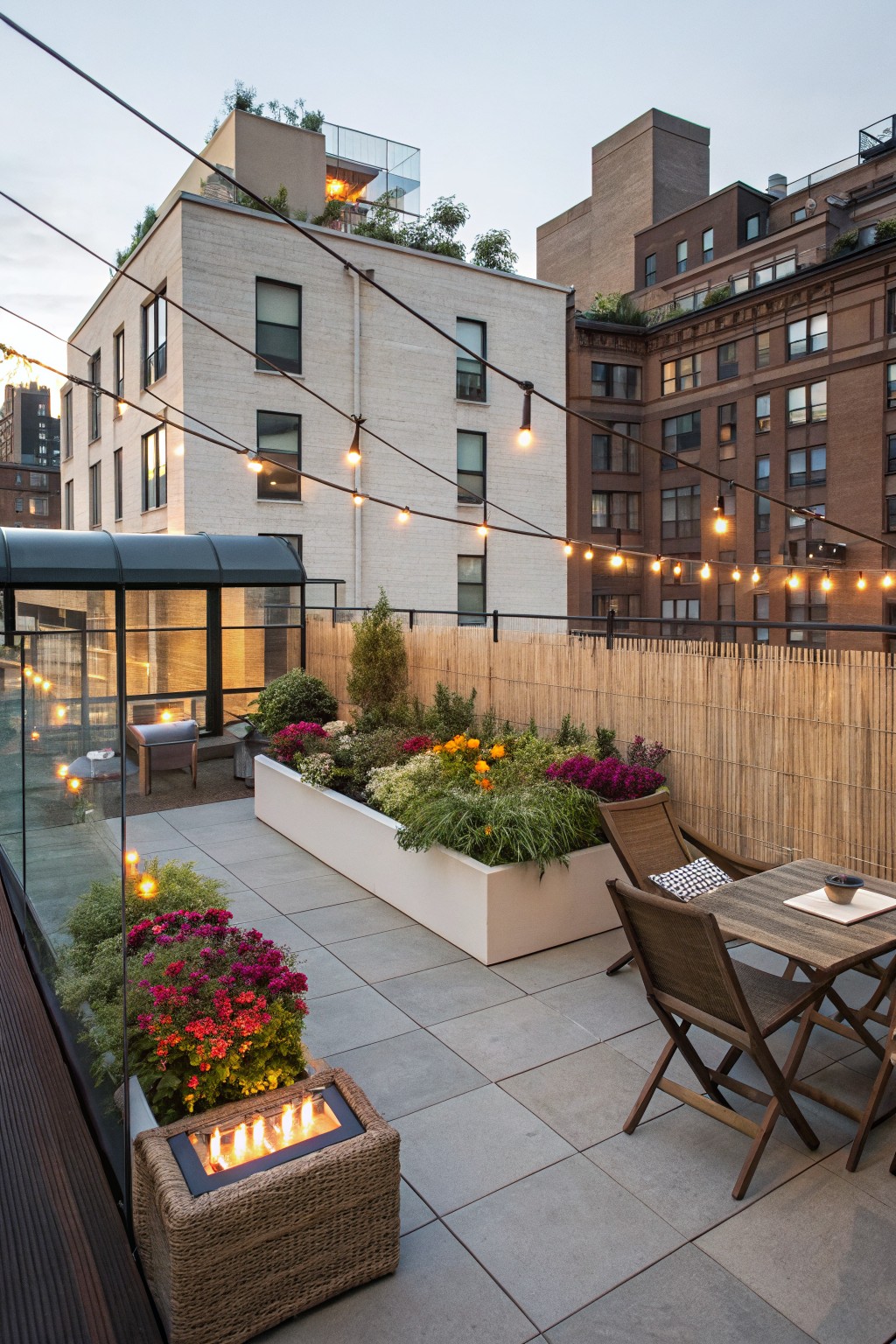 Rooftop terrace with raised white planters filled with colorful flowers like orange zinnias and pink blooms, wooden dining table and chairs, wicker fire pit, string lights, and bamboo fencing amid surrounding city buildings at dusk.