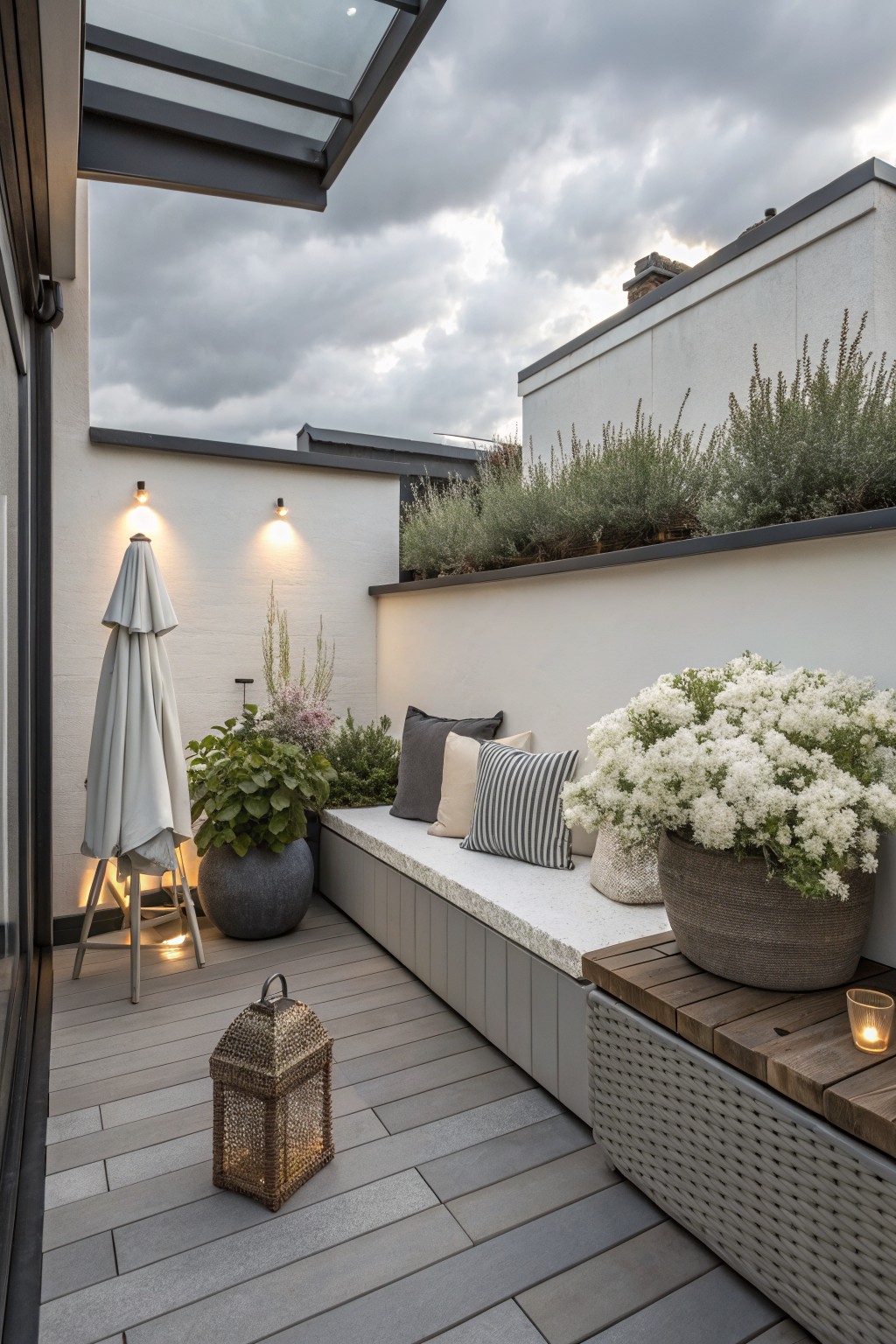 Small rooftop terrace featuring gray plank decking, built-in bench with cushions and wood accent table, large wicker pot of white flowers, potted plants, beige umbrella, lanterns, wall lights, and narrow raised planters with green shrubs atop white walls under a glass canopy.