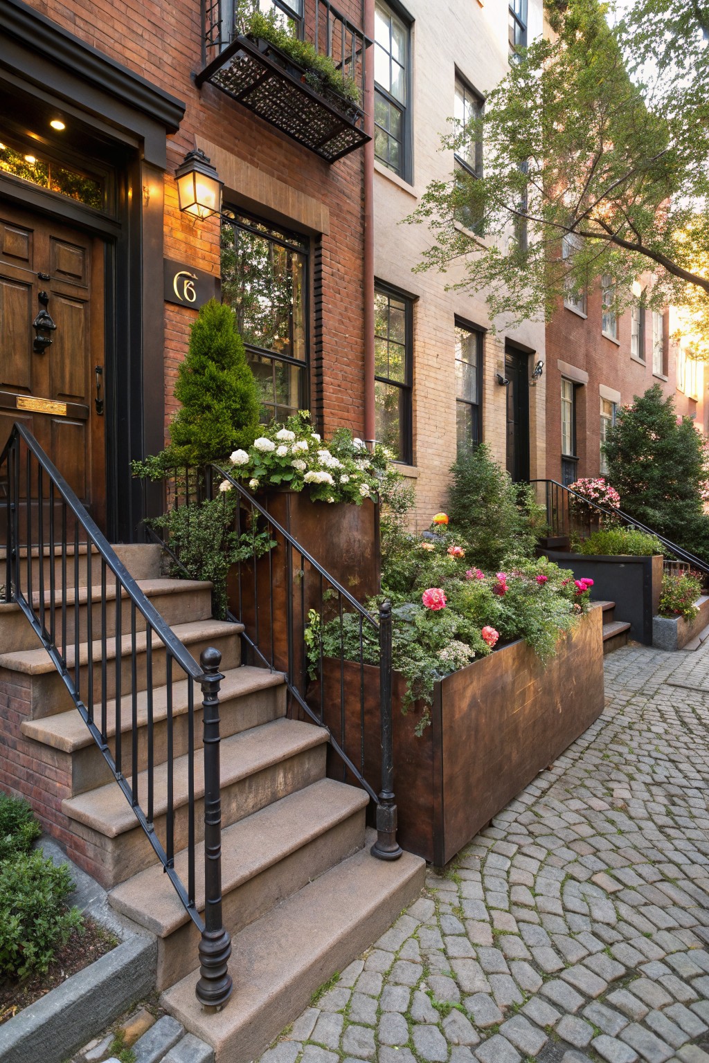 Brick townhouse exterior featuring a wooden front door with number 6, black iron railings along stone steps, and large rectangular copper-colored metal planters filled with pink roses, white hydrangeas, and greenery beside a cobblestone street.