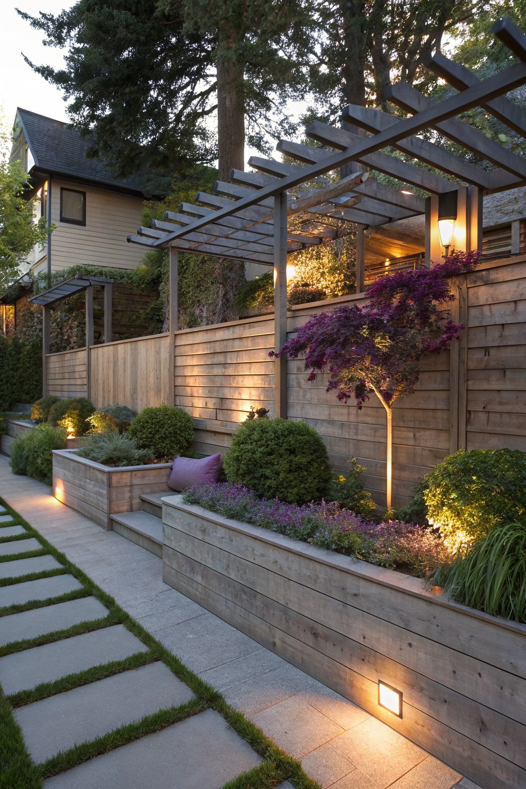 Garden path of stone pavers with grass inlays, bordered by wooden raised planters filled with purple flowers, lavender, and shrubs, next to a tall horizontal wooden plank fence supporting a purple-leaf tree, with ground and wall lights on at evening.