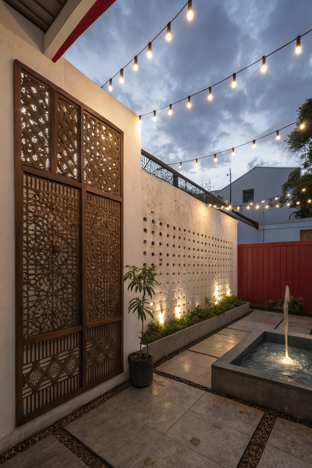 Tall wooden panels with geometric lattice patterns lean against a white textured wall in a backyard patio with string lights overhead, a fountain, plants, and pebble flooring at dusk.