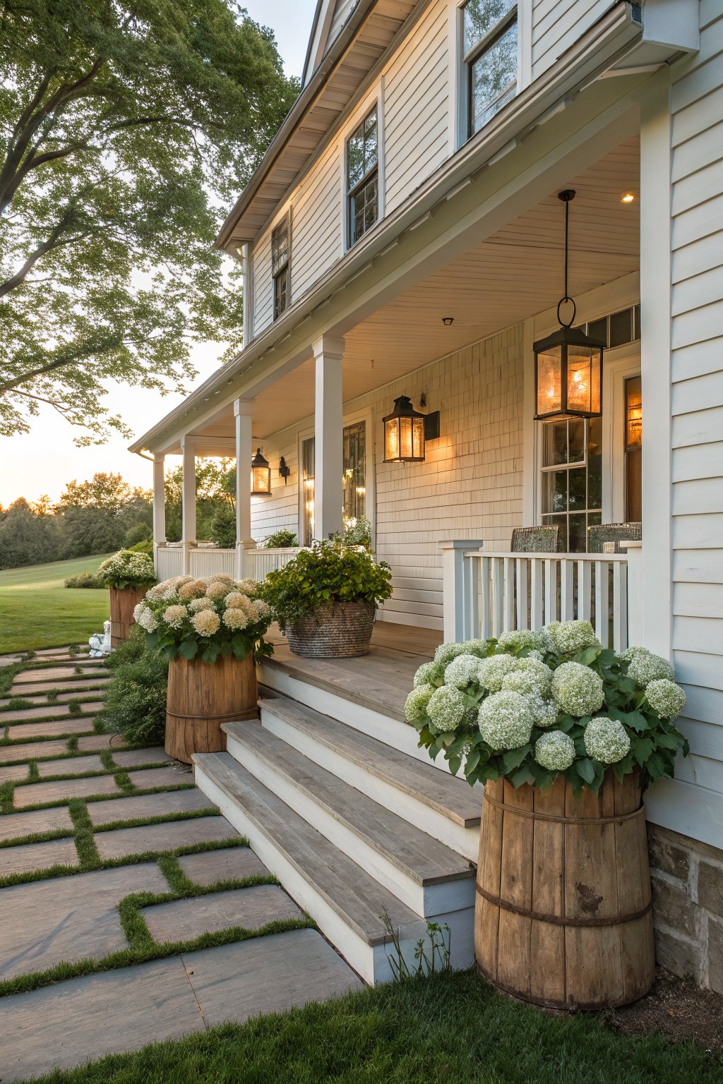 White clapboard house with covered porch and steps, flanked by large white hydrangea plants in wooden barrel planters along a stone path edged with grass, trees and lawn in background.