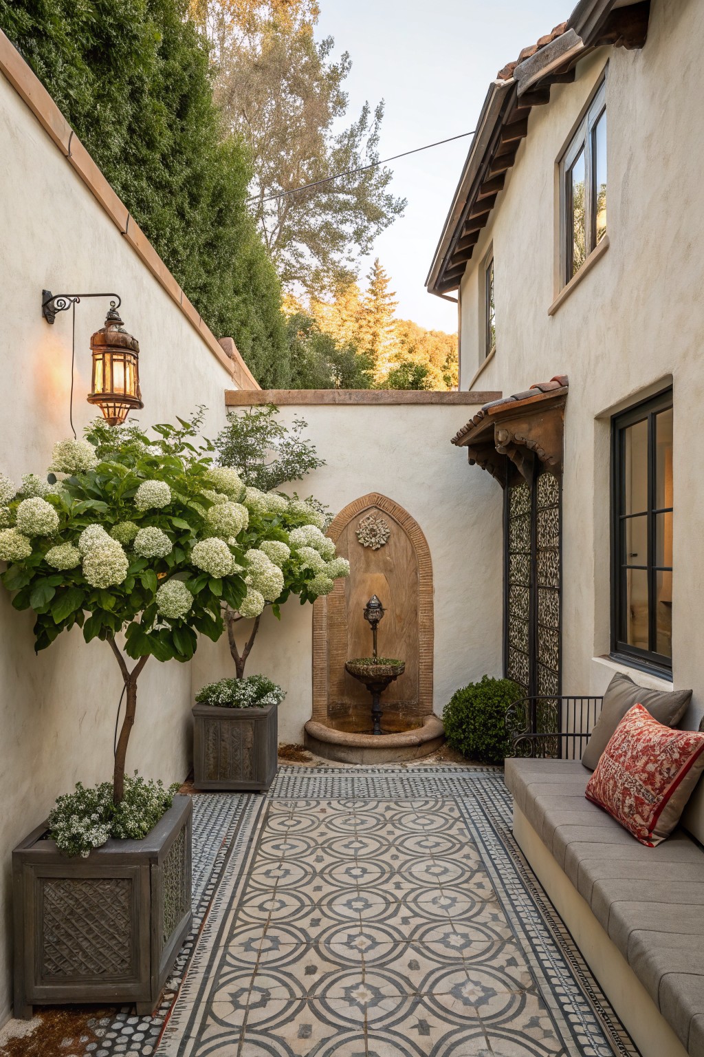 Narrow outdoor pathway between stucco walls featuring large potted hydrangea trees flanking a stone fountain, a tiled floor with geometric pattern, potted plants, a bench with cushions, and a wall lantern.