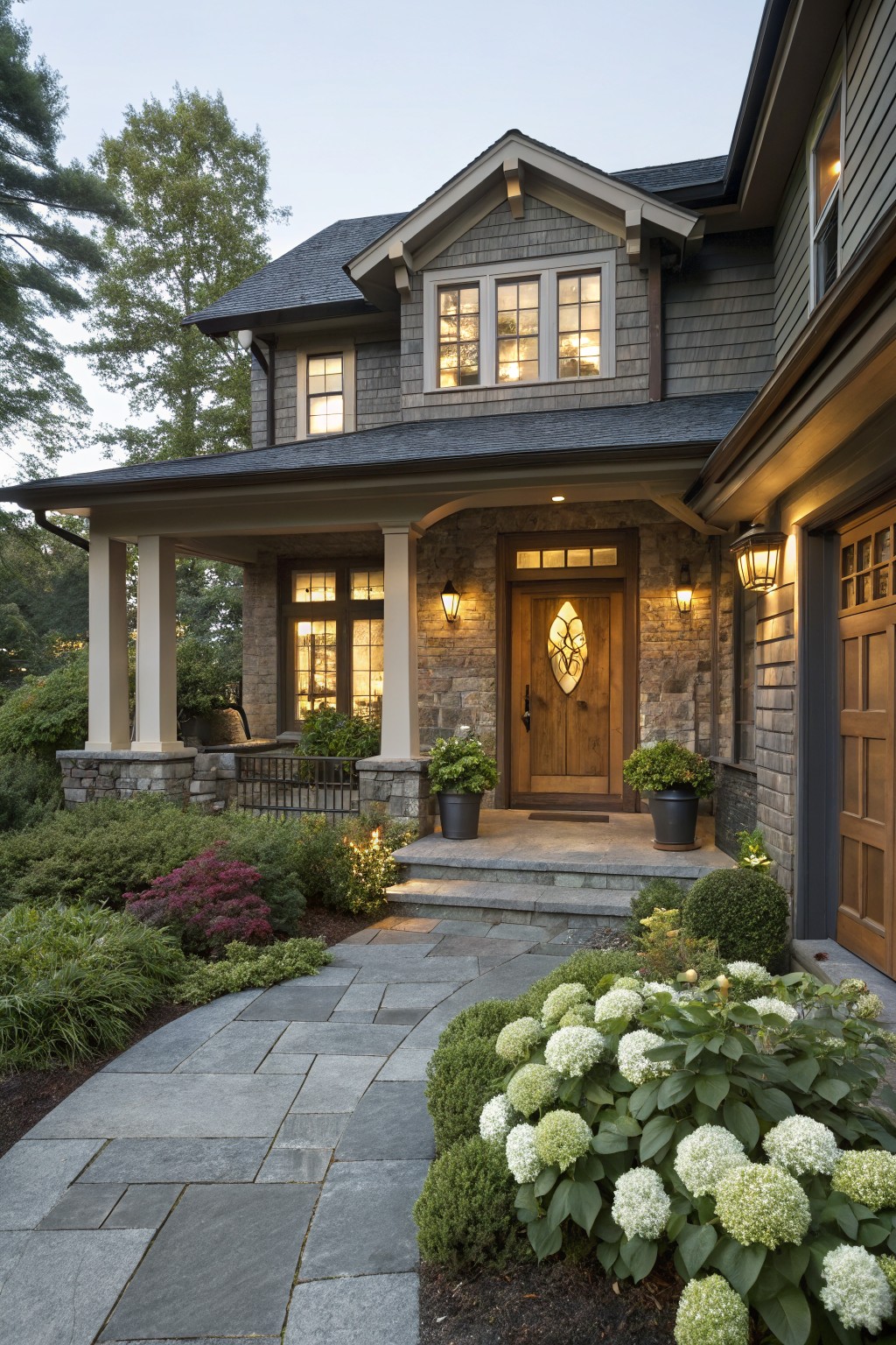 Front view of a shingle and stone house with covered porch, wooden entry door with glass panel, garage door, and a curved gray stone path lined with large white hydrangea bushes, shrubs, and perennials leading to porch steps.