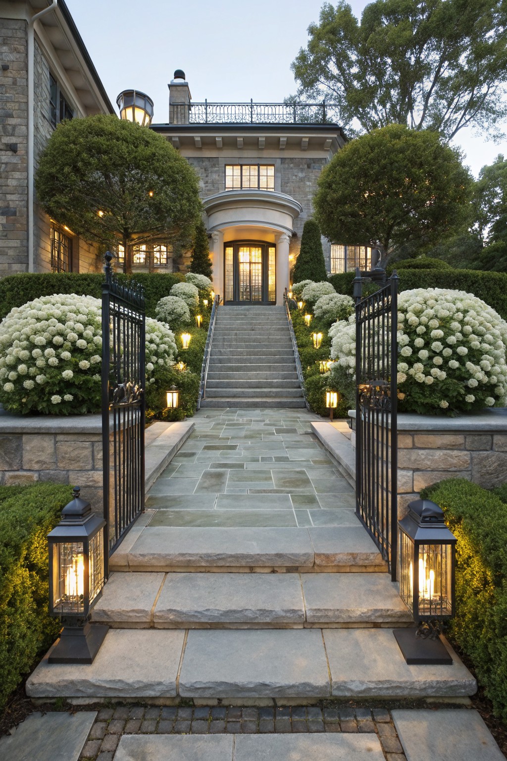 Stone pathway leading to a wrought iron gate at a large house entrance, flanked by large white hydrangea bushes and boxwood hedges, with stone steps, lanterns, and evening lighting.