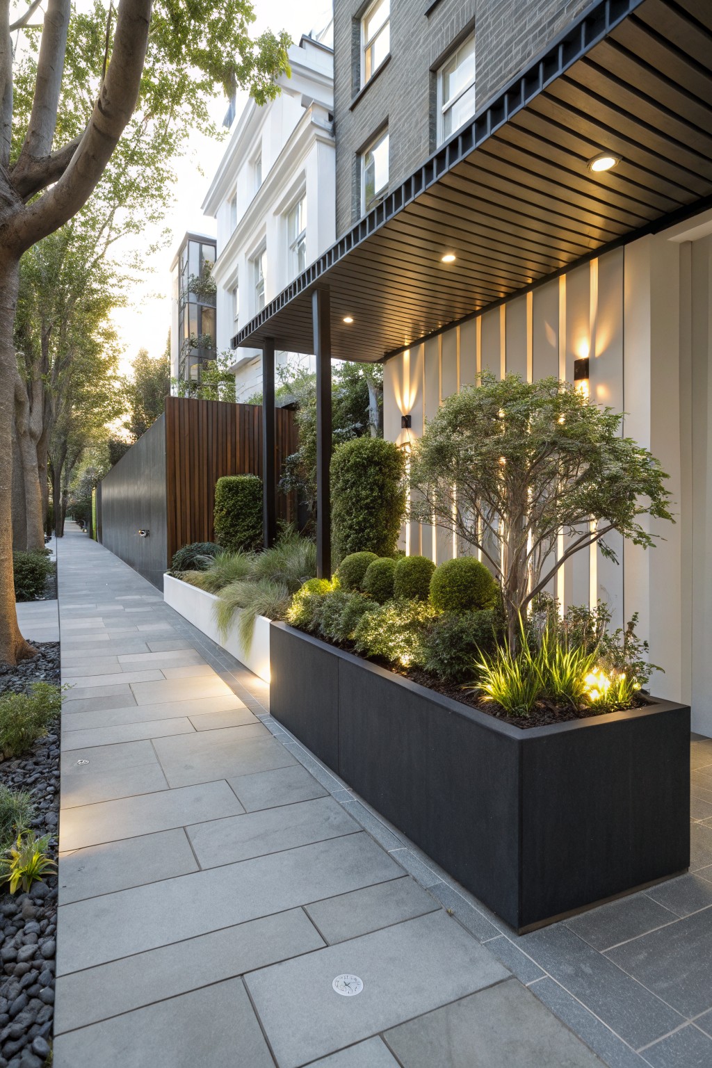 Narrow gray stone walkway bordered by long black rectangular planters filled with ornamental grasses and shrubs, with trees and brick buildings nearby.