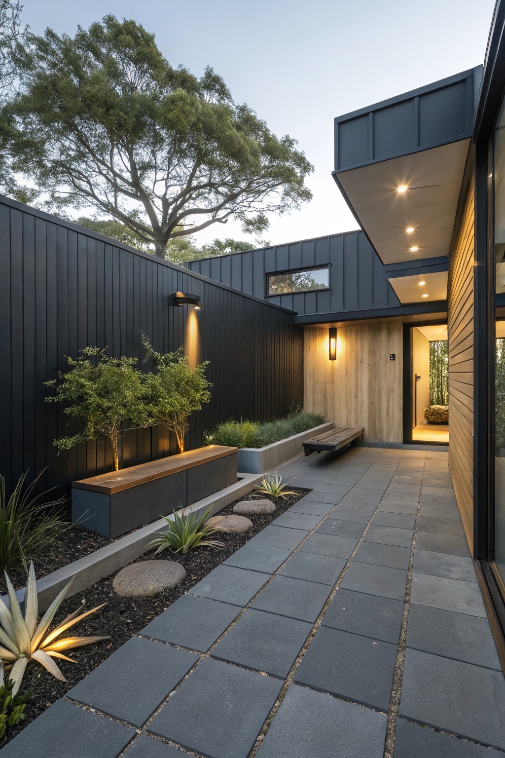 Dark gray square paver pathway leading to a modern timber entry door, edged with agave plants and grasses, a timber bench planter, black vertical cladding fence, and wall-mounted lights.