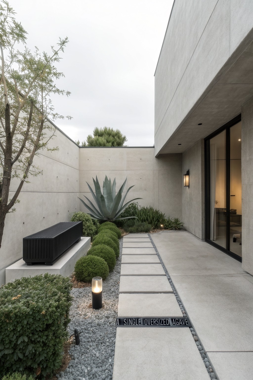Minimalist landscape beside a concrete modern house featuring a large single agave plant, rounded boxwood shrubs, a stone paver path with gravel edges, black bench, and glass entry doors.
