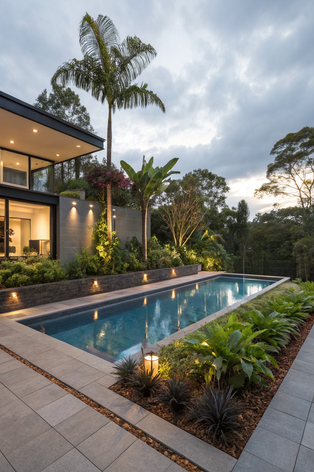 Contemporary house exterior at dusk with infinity-edge pool, gray stone edging and retaining walls around tropical garden beds, pathway, and lighting.