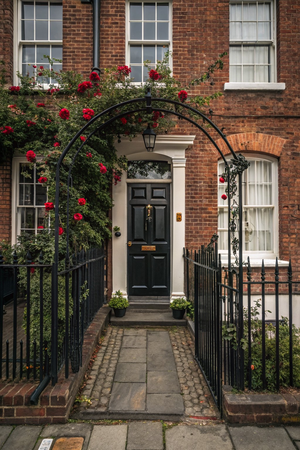 Red brick townhouse exterior featuring a black front door, black wrought iron fence and arched trellis covered in red climbing roses over the front path.