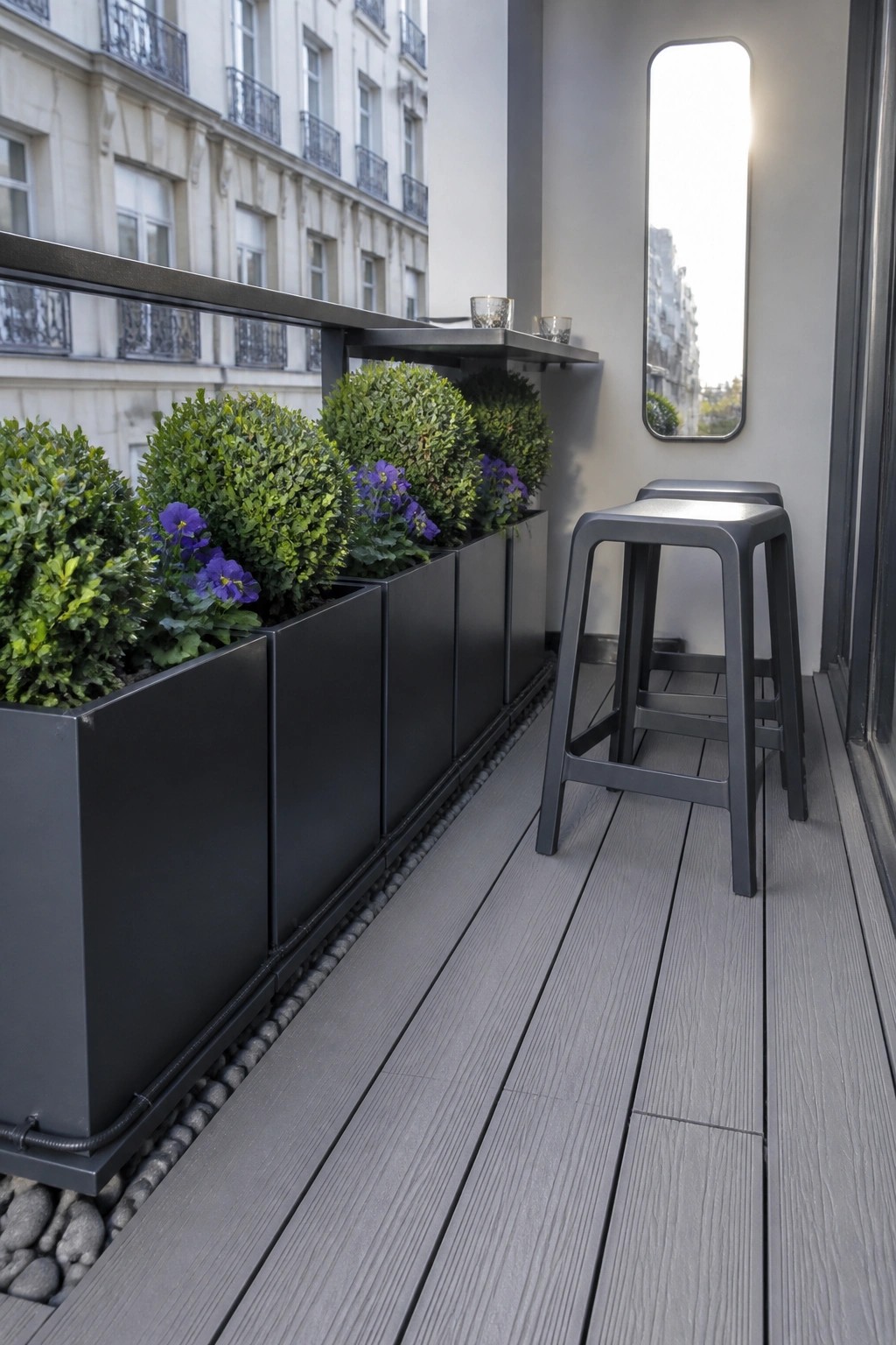 A balcony with a row of black rectangular planters holding green shrubs and purple flowers along the edge, gray decking, and two dark stools.