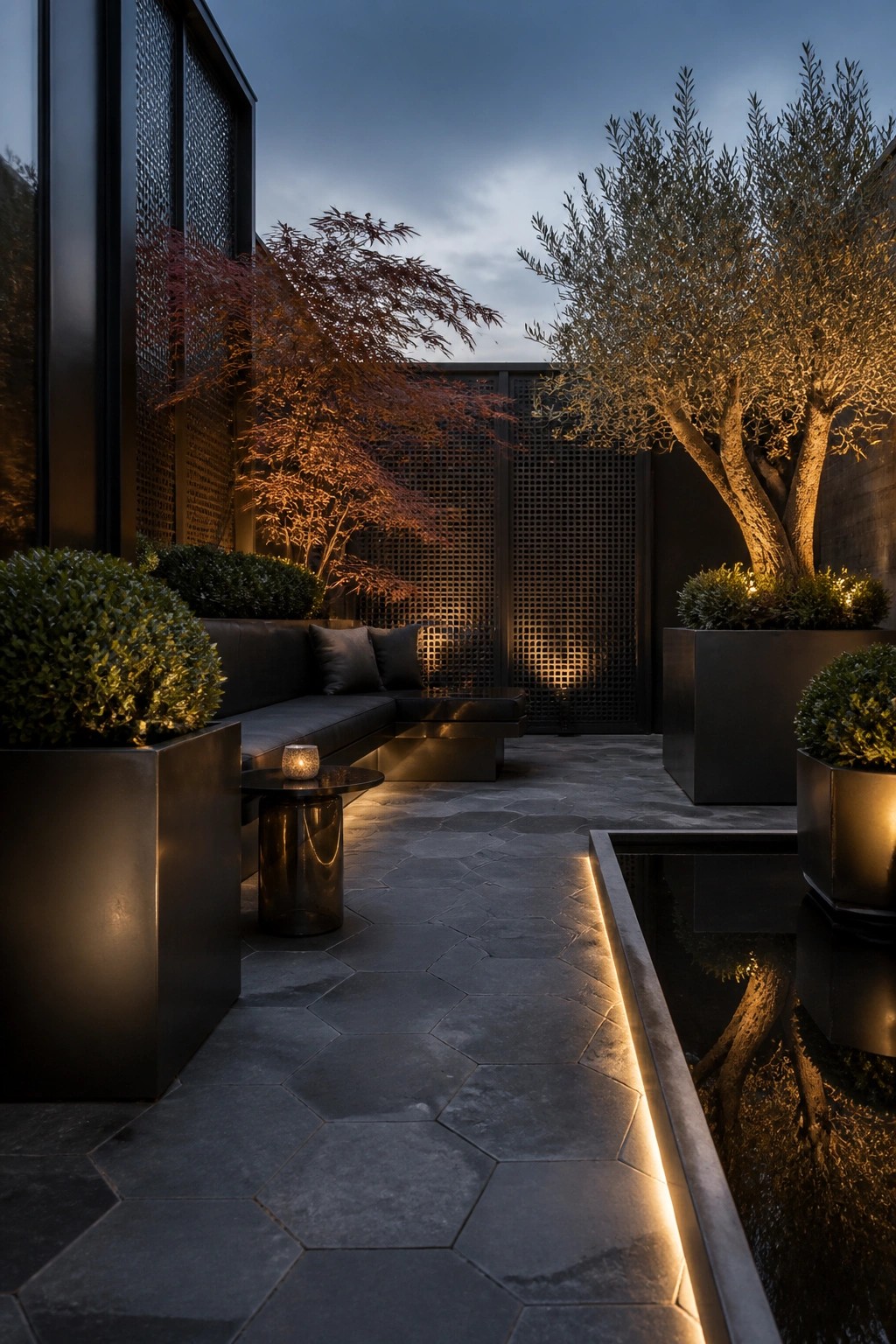 An evening view of a modern outdoor patio with dark sectional seating, large metal planters holding trees and shrubs, a narrow water feature, and lattice screens along the back wall.